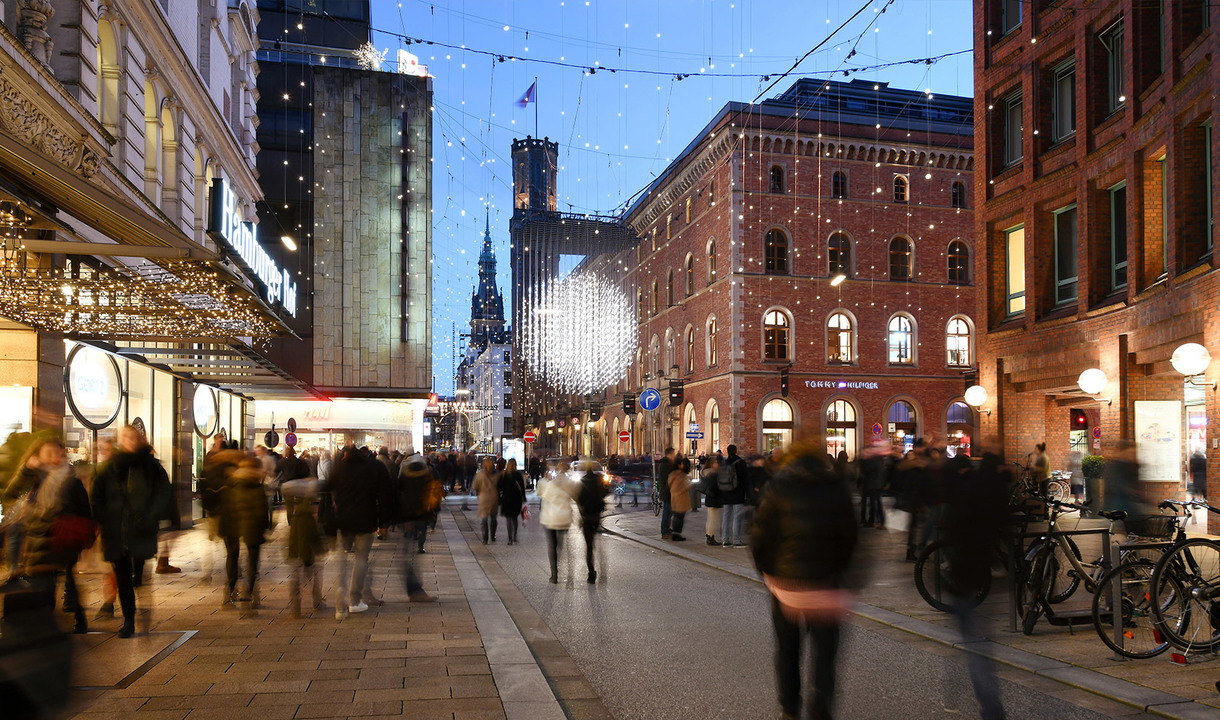 Affollata strada pedonale al crepuscolo con calde luci festive, vetrine illuminate e una grande installazione luminosa sopra la folla