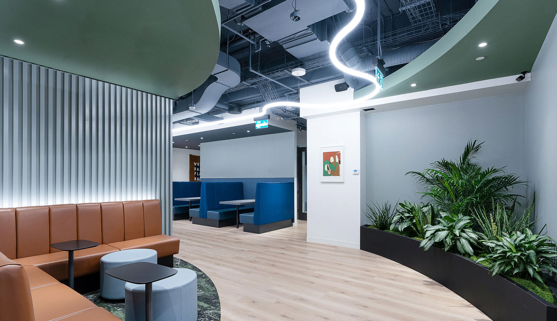 Lounge ufficio moderno con luce a soffitto LED sinuosa, faretti incassati, sedute in pelle, cabine blu e piante da interno.