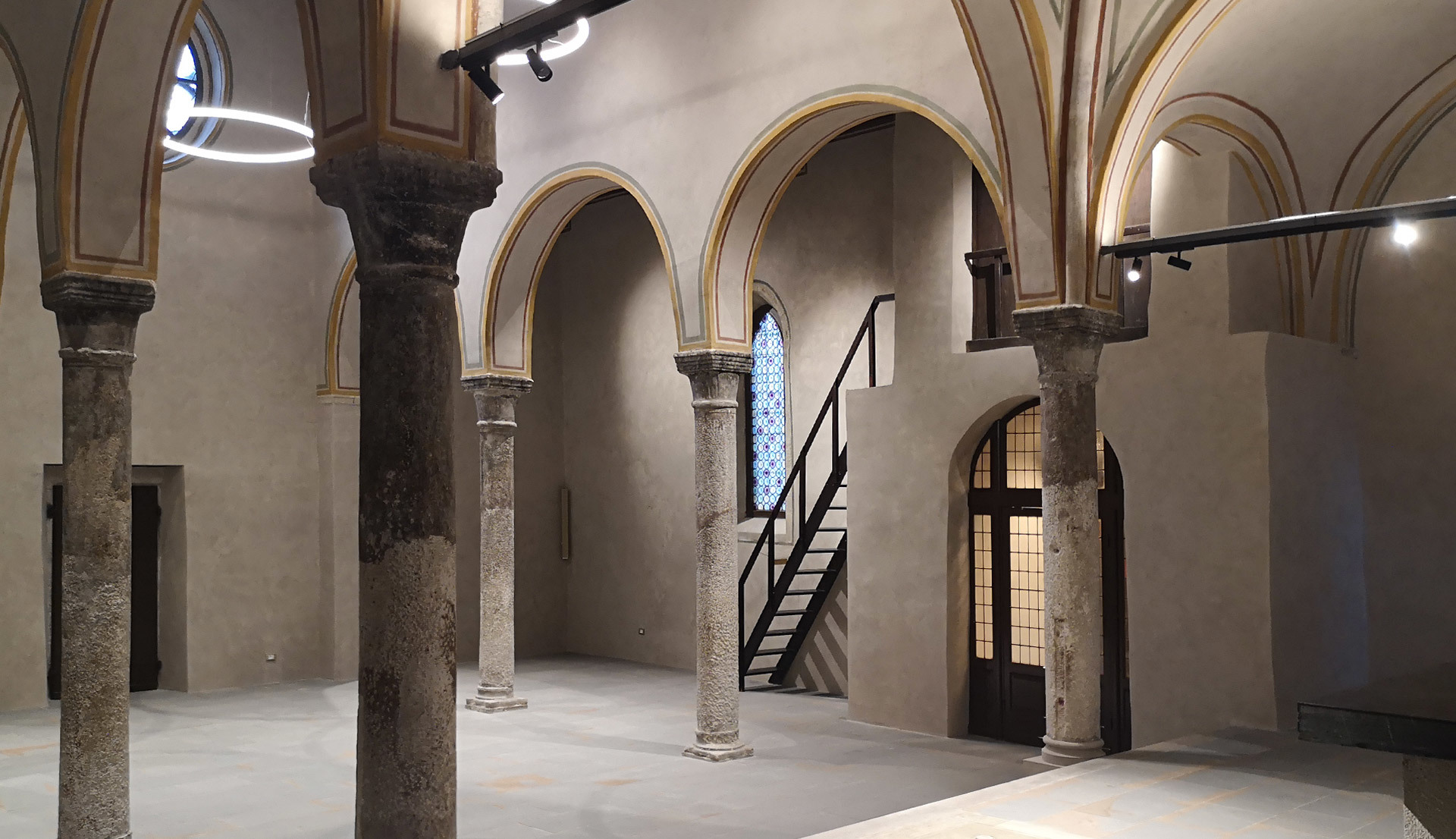 Sala storica restaurata con archi e colonne in pietra, illuminata da sospensione circolare e faretti su binario Artemide.