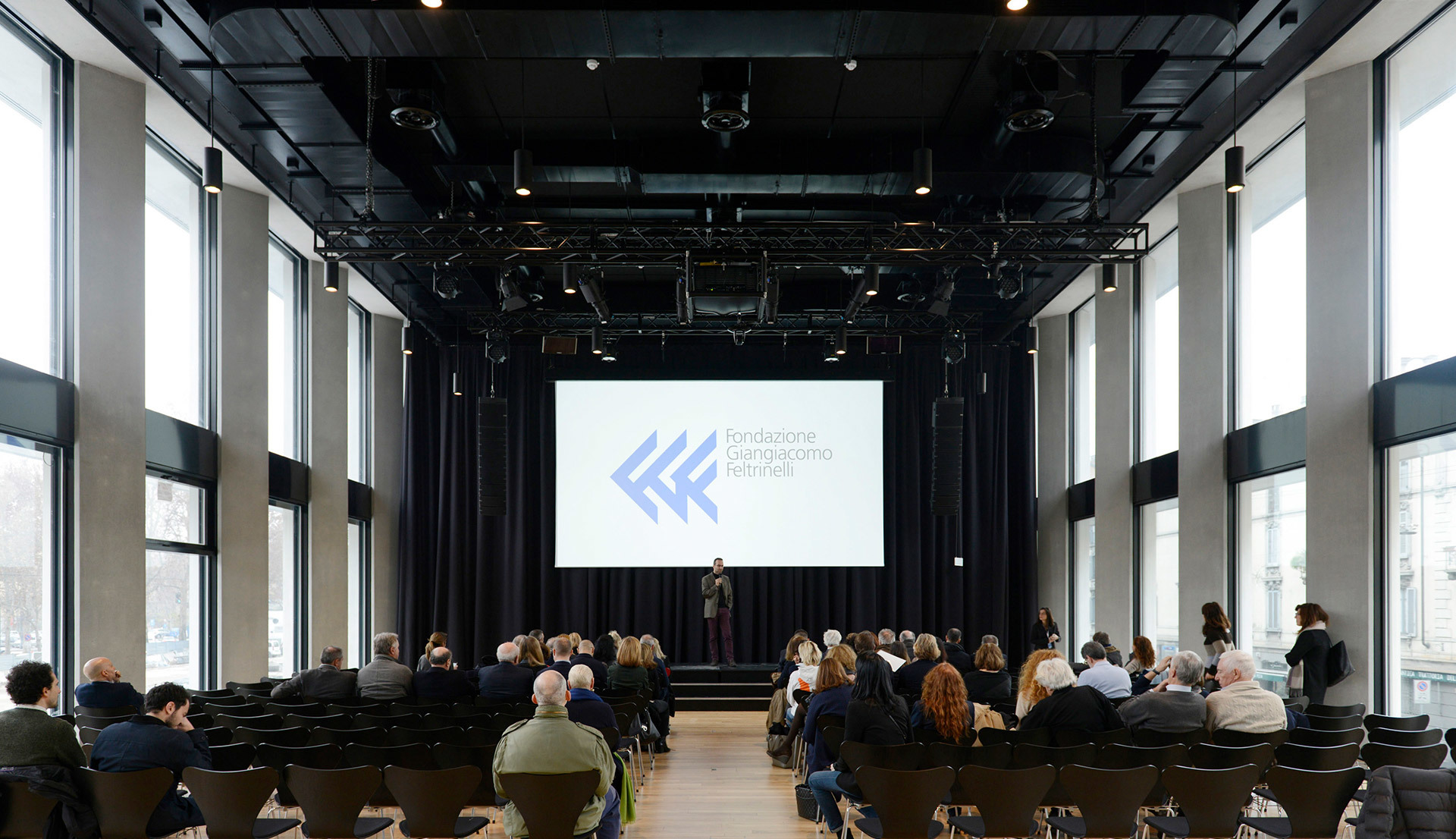 Sala conferenze con pubblico seduto di fronte a un relatore sul palco, grande schermo di proiezione, alte finestre e illuminazione industriale a soffitto.