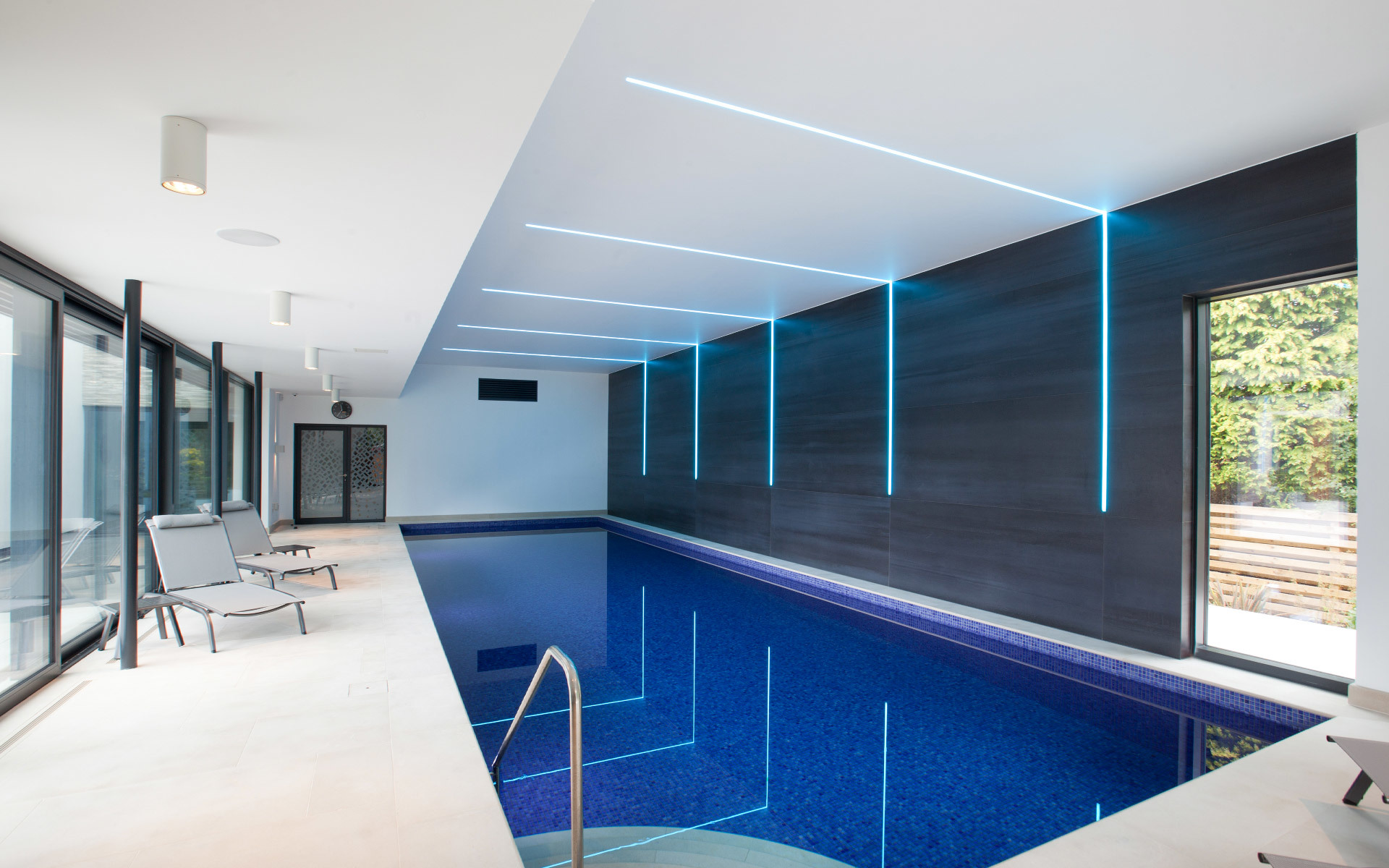 Indoor pool with blue mosaic tiles, illuminated by cyan linear LED lights on ceiling and dark accent wall, with lounge chairs.
