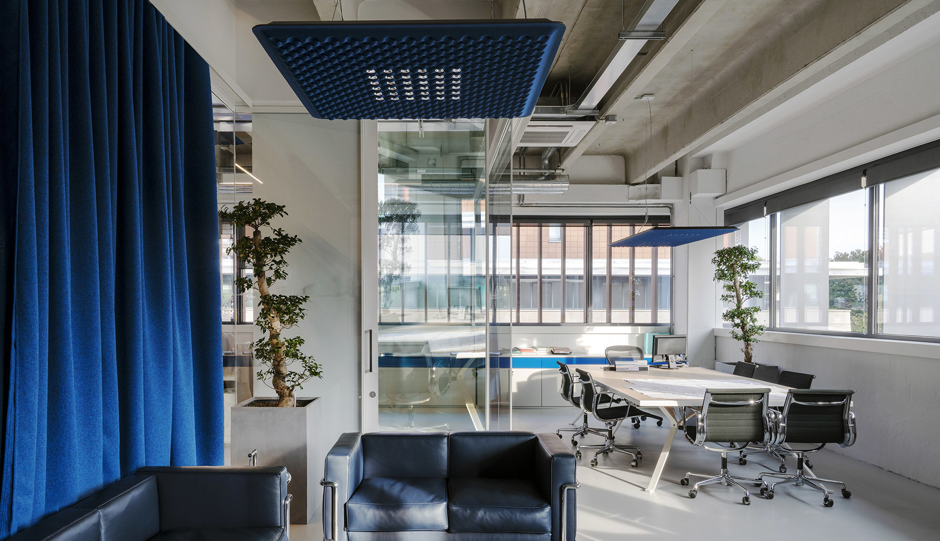 Ufficio moderno con lampade a sospensione Artemide blu sopra un tavolo riunioni, divano lounge, pareti in vetro e bonsai.