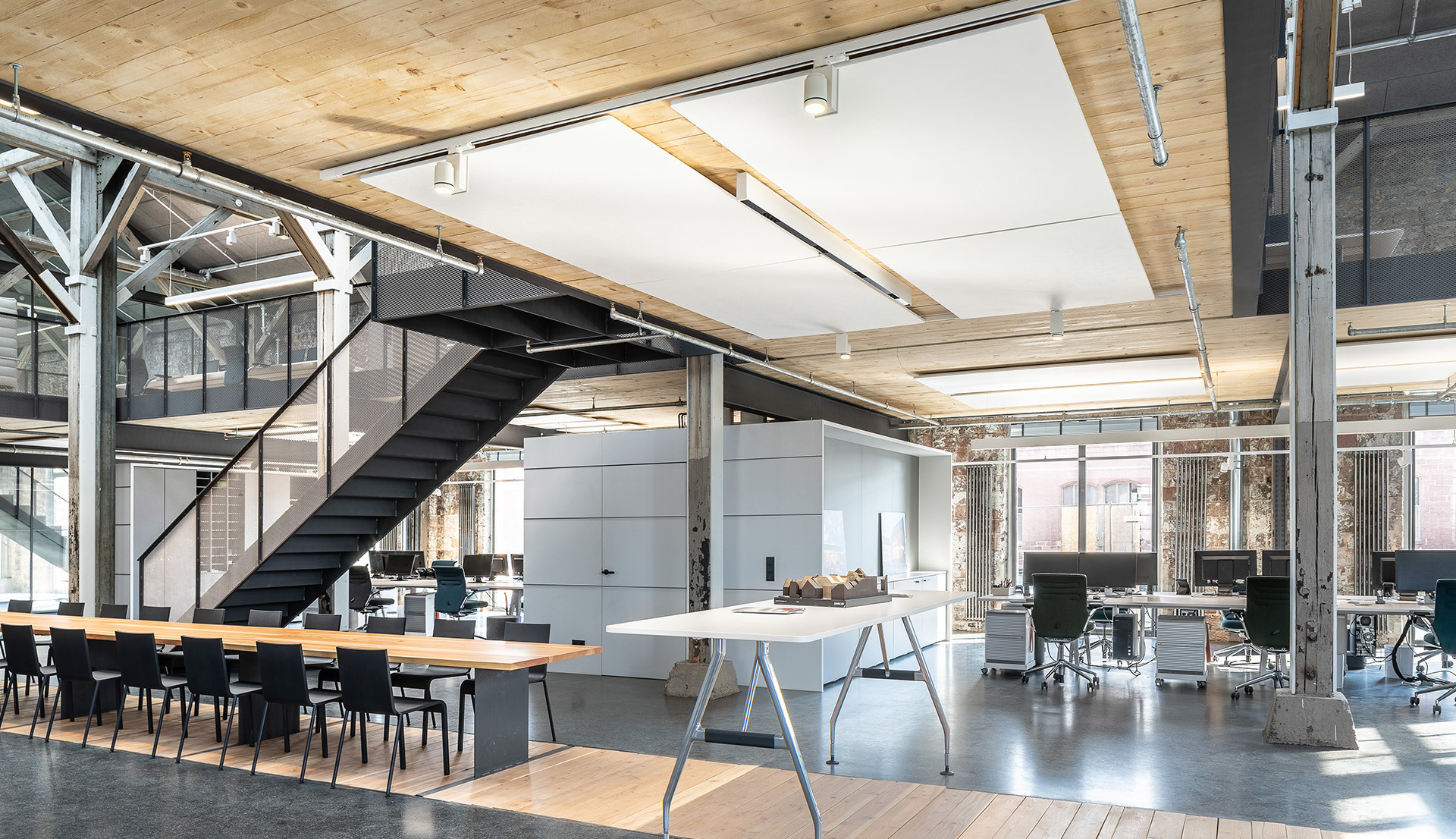 Ufficio open space moderno con luci a soffitto Artemide, travi in legno, pavimenti in cemento, postazioni di lavoro e scala industriale in acciaio.