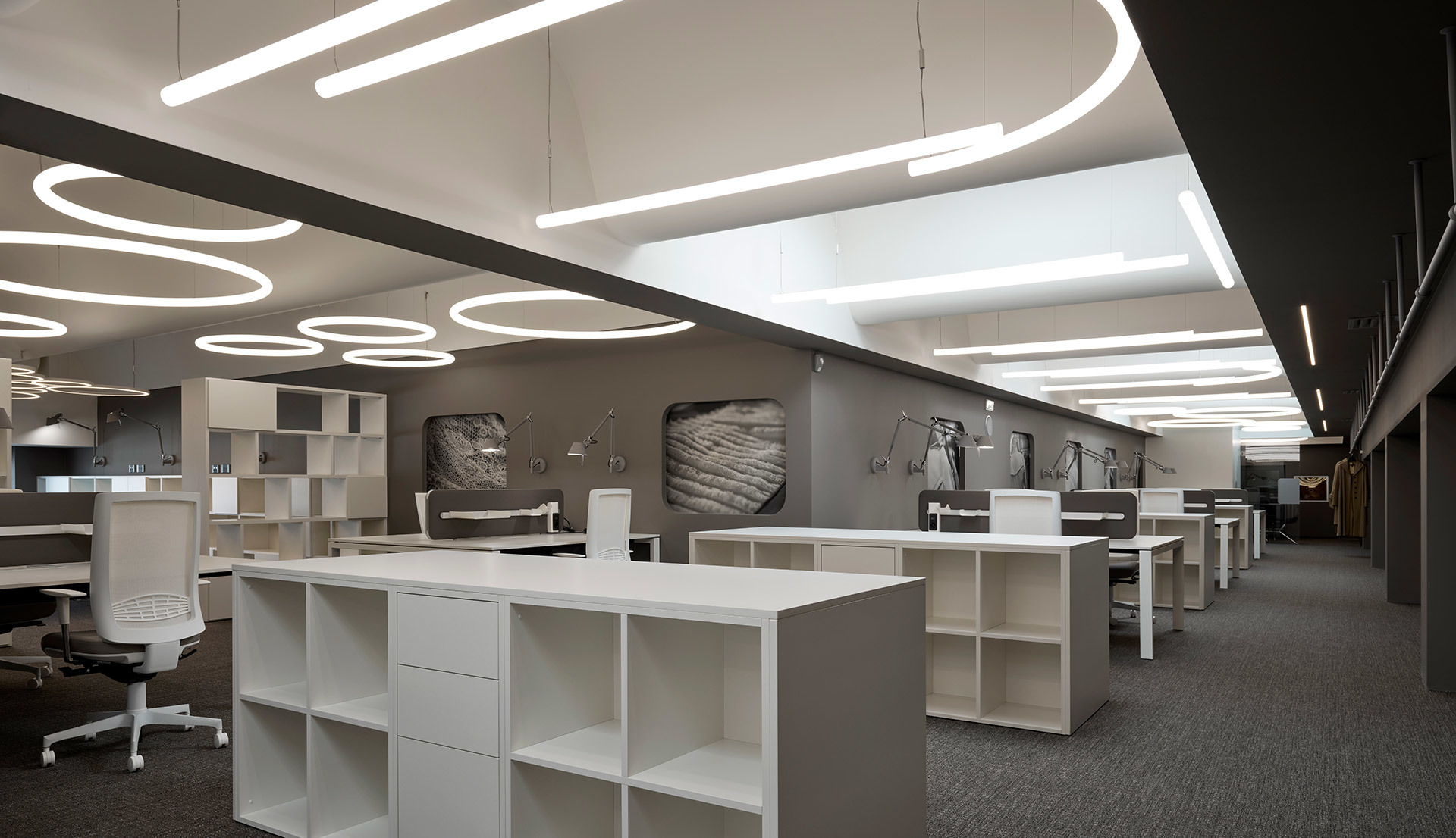 Showroom ufficio moderno con lampade a sospensione circolari e plafoniere lineari Artemide che illuminano uno spazio aperto con scaffalature bianche.