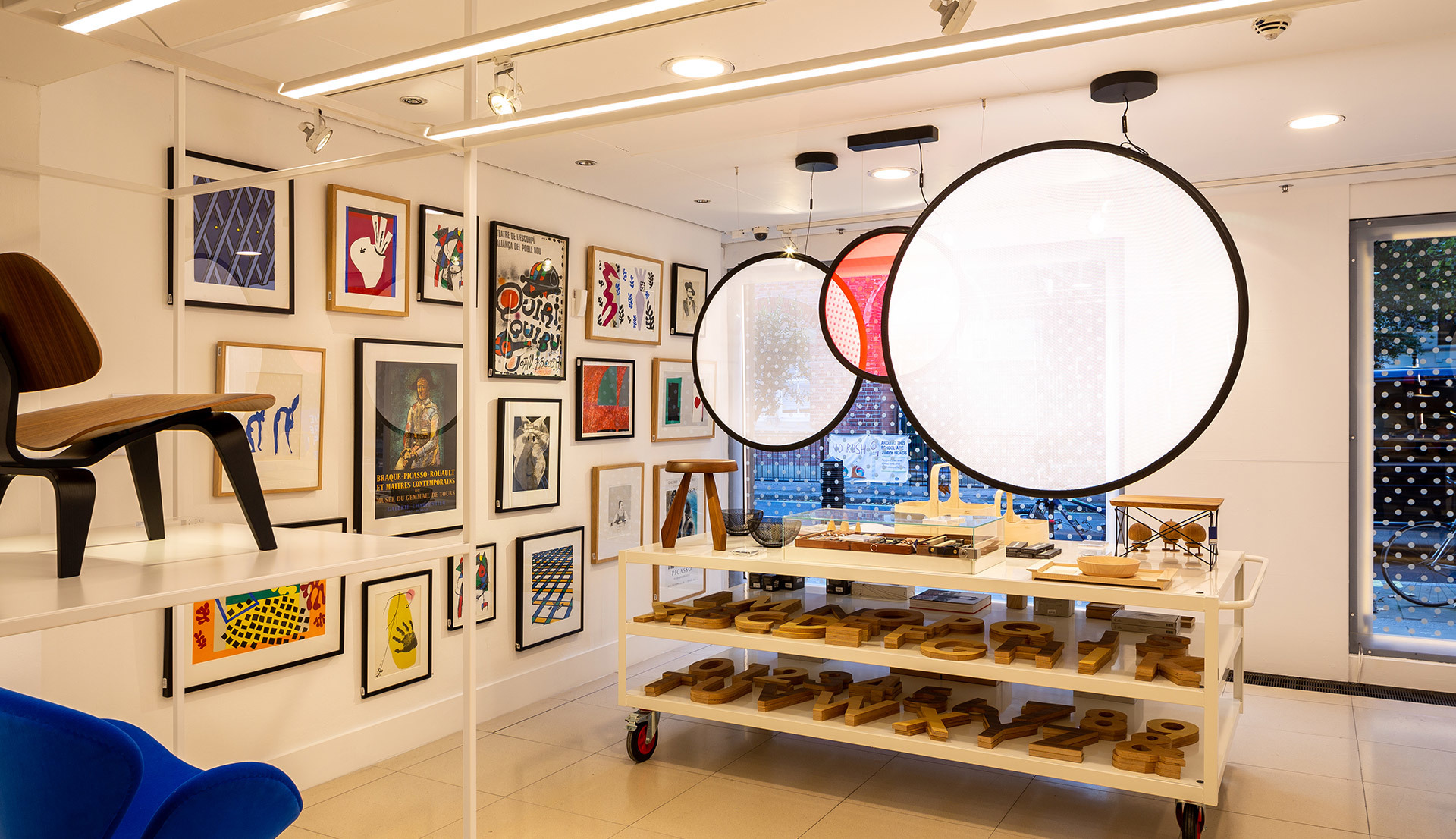 Spazio commerciale con grandi lampade a sospensione circolari con cornici nere, parete galleria con opere incorniciate e carrello espositivo con oggetti in legno.