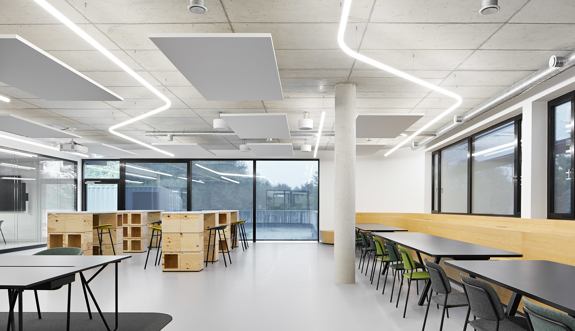 Spazio di lavoro aperto moderno con luci a soffitto LED lineari geometriche, pannelli acustici, mobili modulari in legno e lunghi tavoli con sedie verdi.