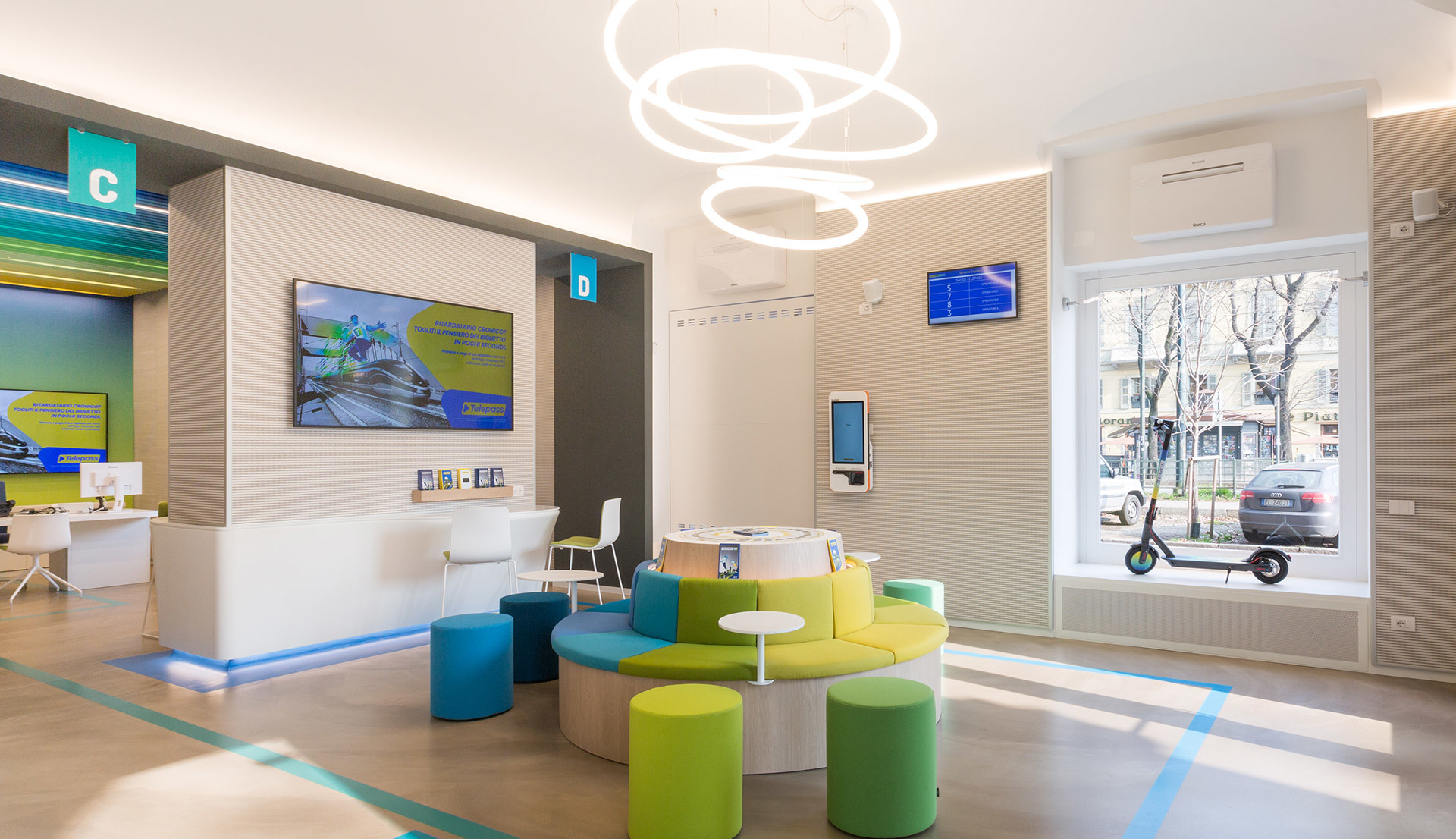 Lounge ufficio moderno con lampade a sospensione ad anelli circolari Artemide, sedute colorate, reception e schermi digitali.