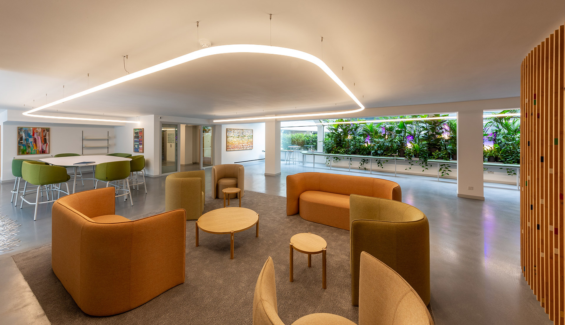 Modern lounge with curved LED ceiling light, warm orange sofas, wooden tables, and a lush green wall visible through floor-to-ceiling windows