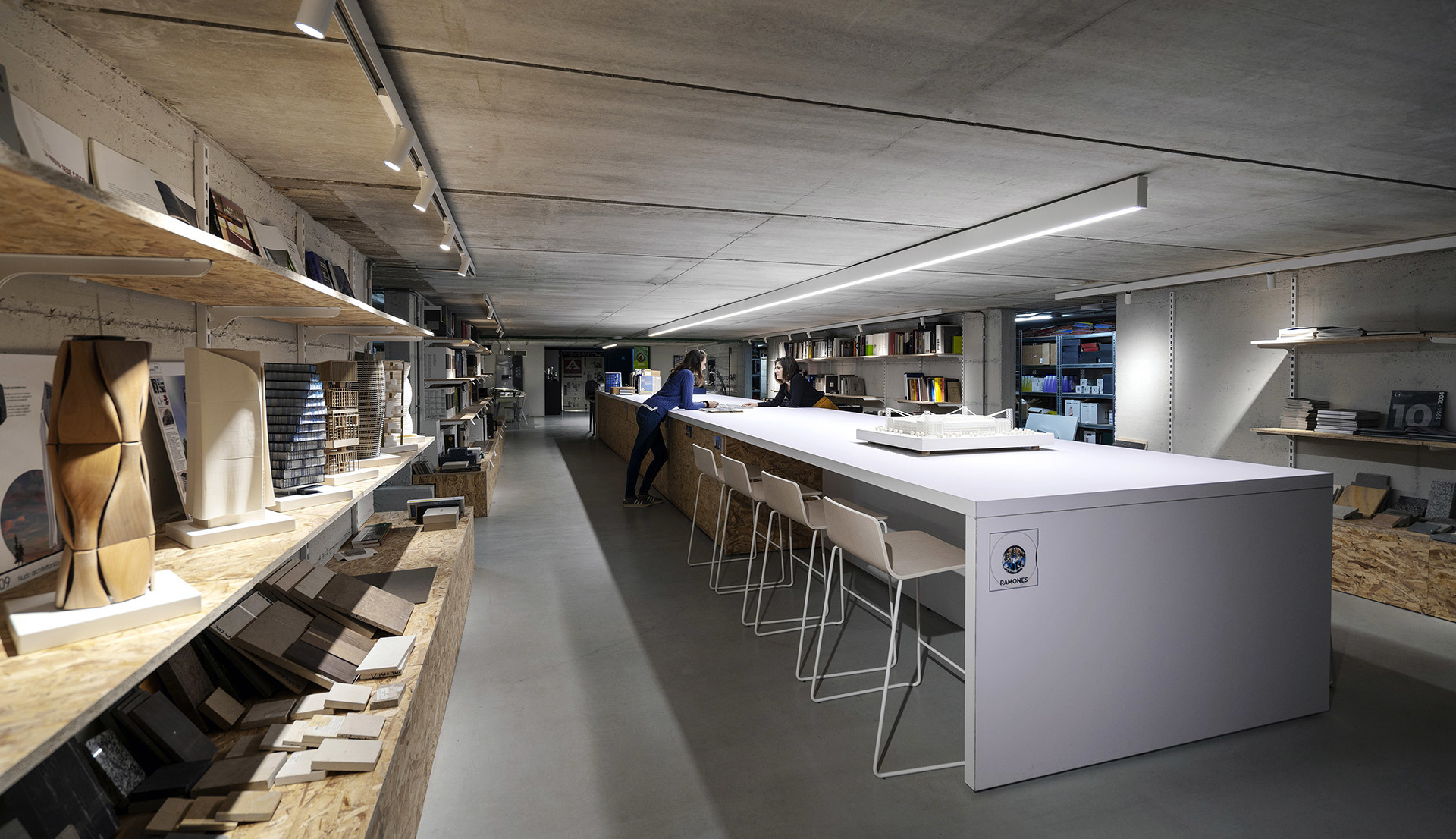 Spazio di lavoro moderno con lungo tavolo bianco, sgabelli, scaffali con campioni e illuminazione a binario LED lineare Artemide su soffitto in cemento.