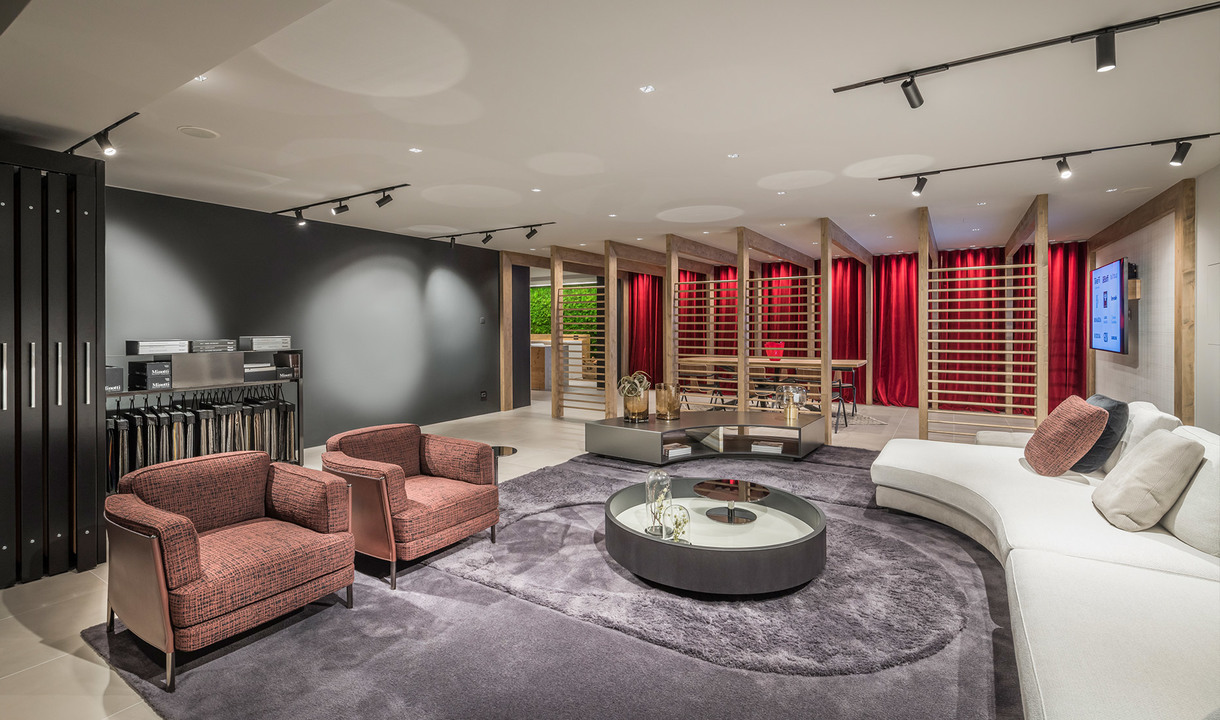 Salotto elegante con faretti su binario a soffitto, poltrone rosa, divano curvo bianco, tavolino rotondo su tappeto grigio, tende rosse sullo sfondo