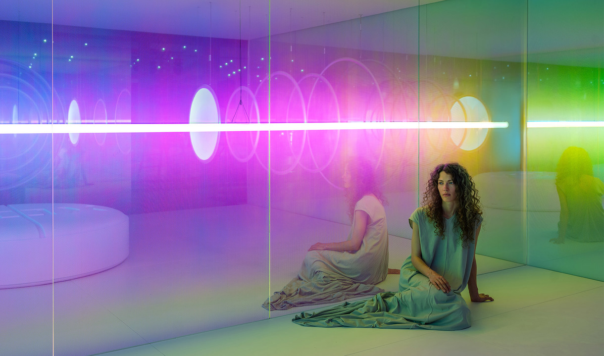 Donna seduta in una stanza immersiva immersa in una luce vibrante rosa, viola e verde proveniente da un'installazione di tubi al neon orizzontali