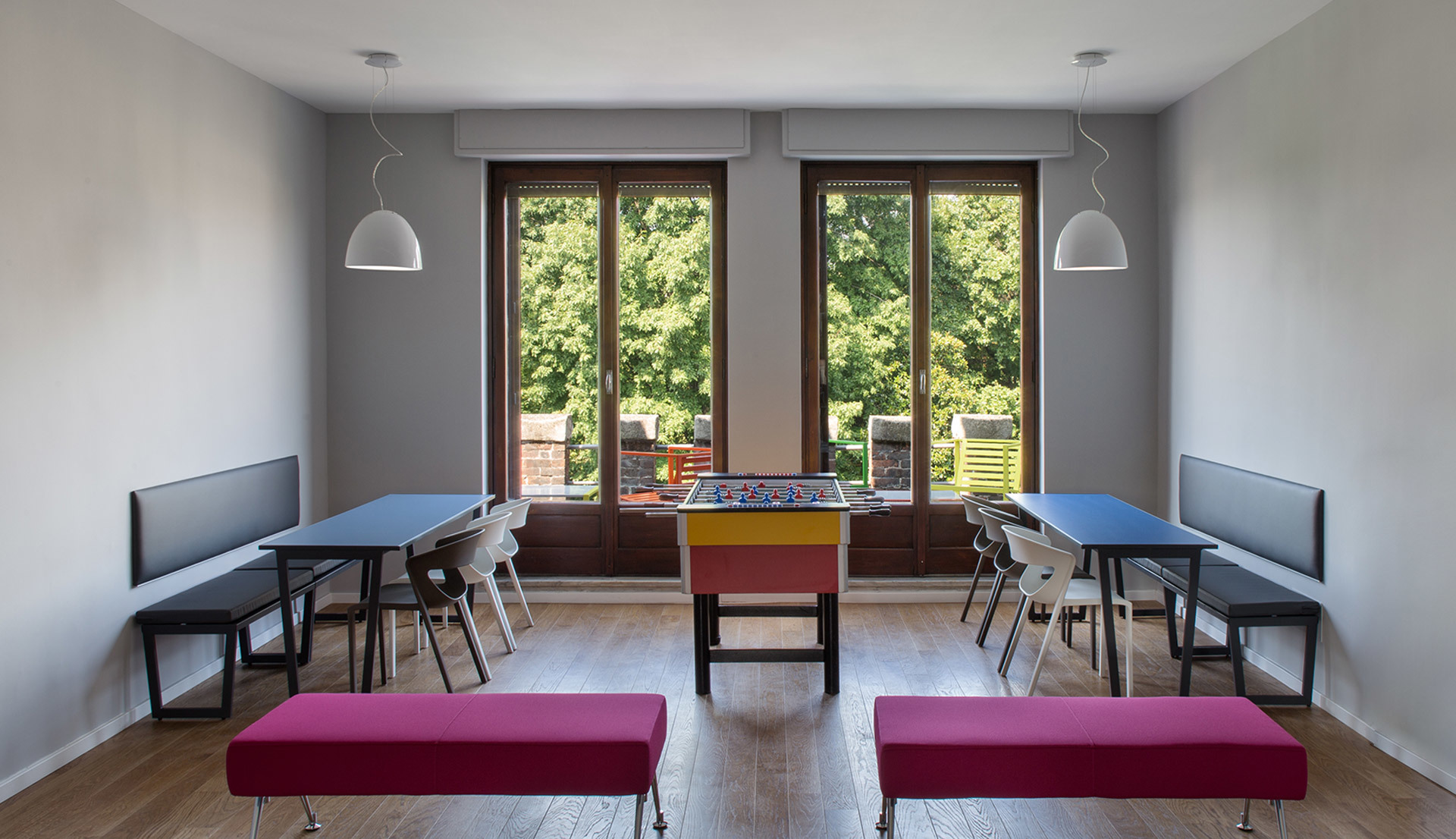 Playroom with two Artemide white pendant lamps over blue tables, magenta benches, and a foosball table near large windows.