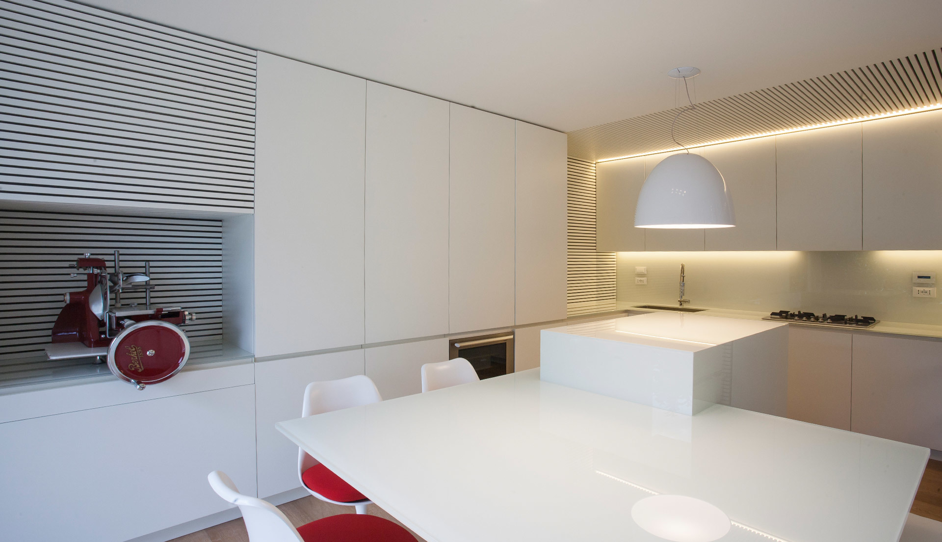Modern white kitchen with large pendant lamp and warm LED strip lighting above cabinets, featuring a red meat slicer accent.