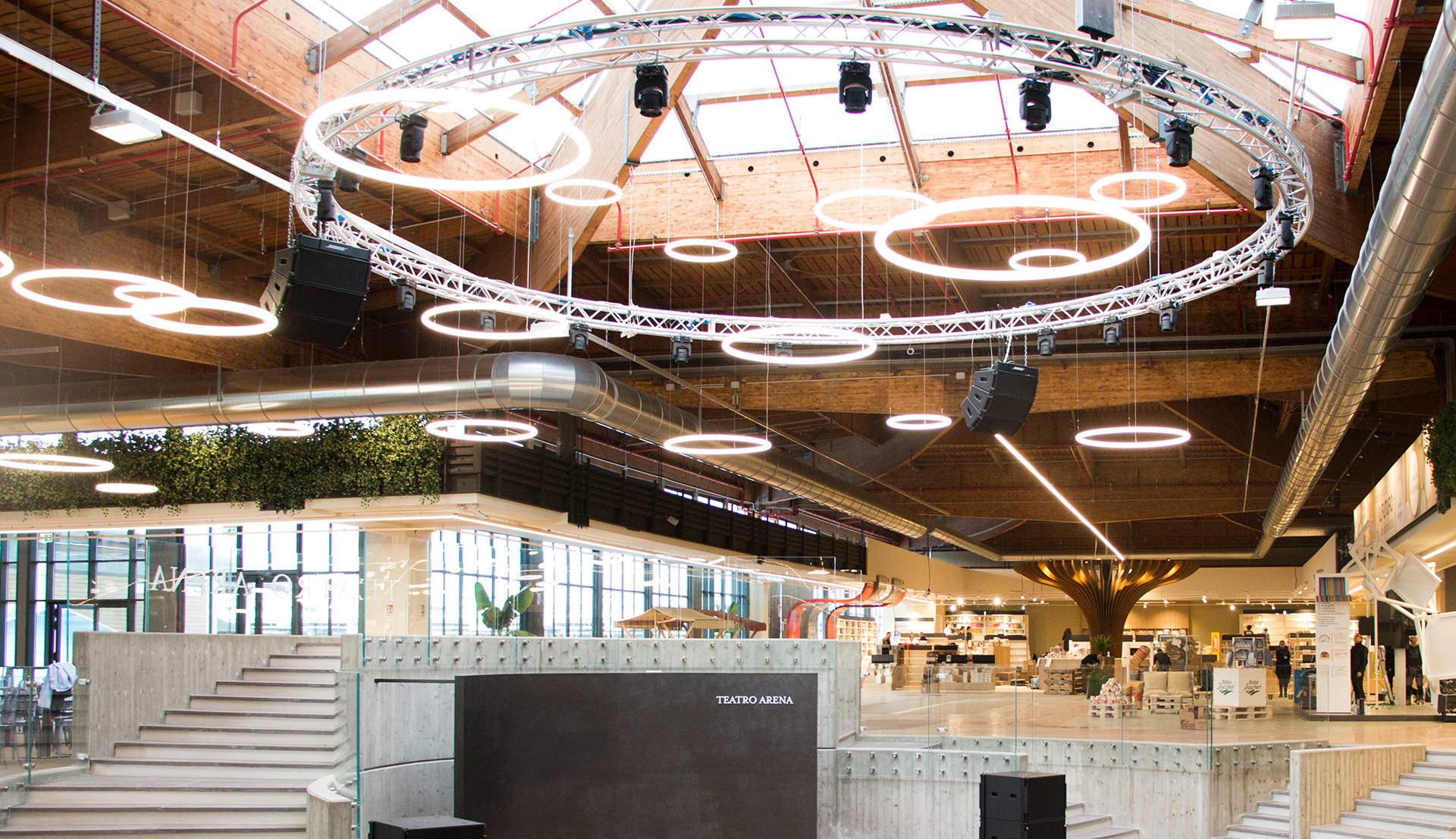 Large modern atrium with circular LED ring pendant lights suspended from brick ceiling above a central stage and retail area