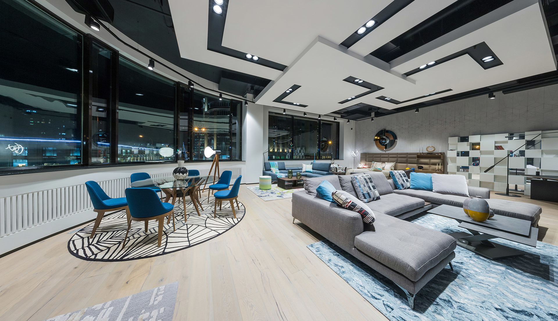 Moderno spazio living open space con luci a soffitto incassate, grande divano componibile grigio, sedie da pranzo blu e finestre a tutta altezza di notte
