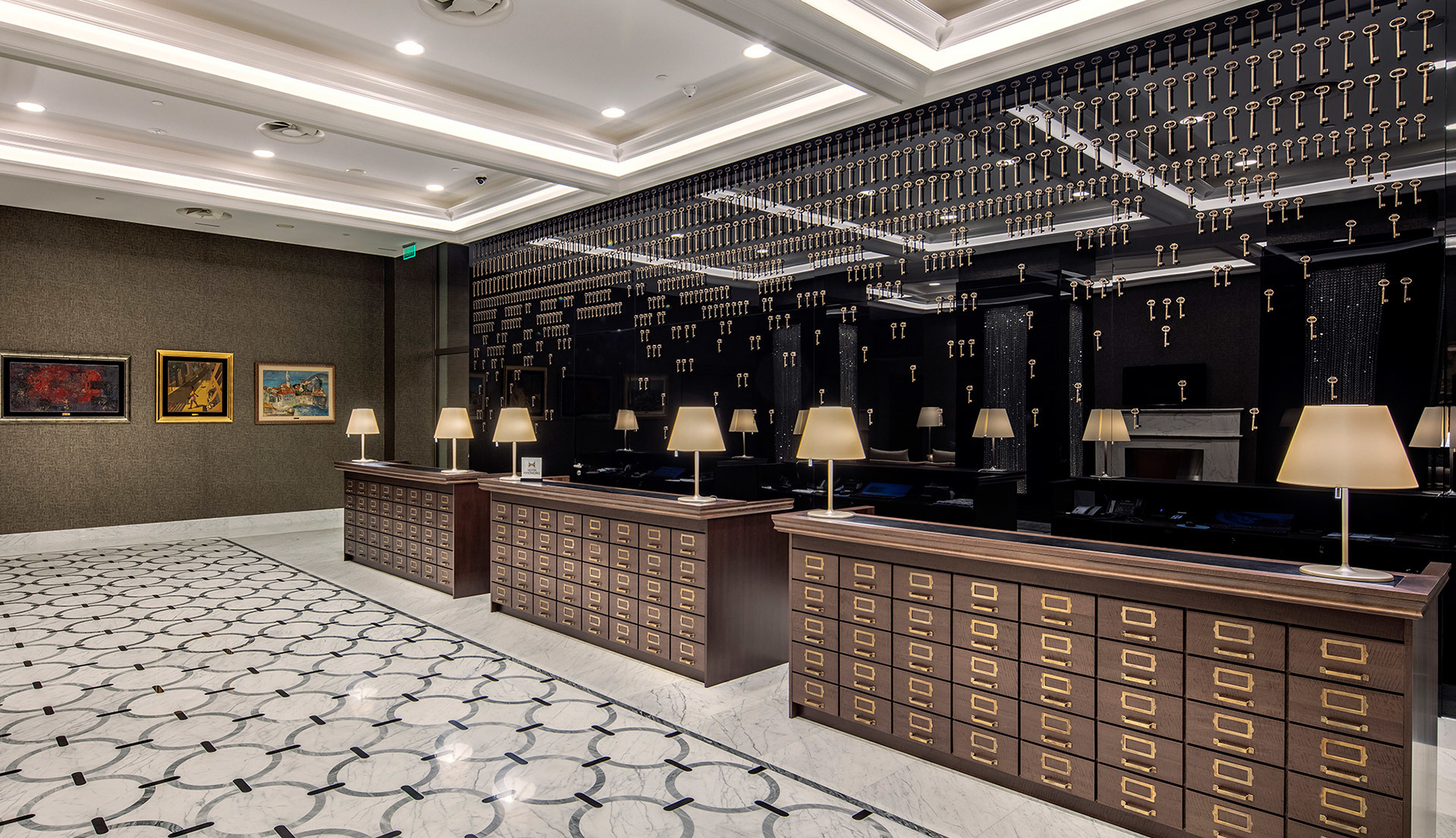 Elegante reception d'albergo con banconi in legno scuro, lampade da tavolo con paralumi color crema, parete porta-chiavi e illuminazione a soffitto incassata.