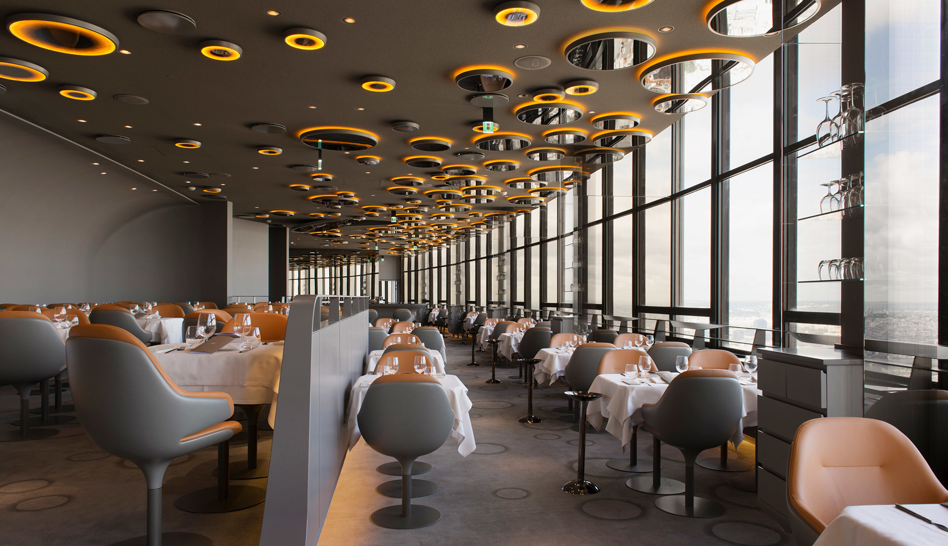 Soffitto di ristorante elegante con multiple lampade a sospensione circolari Artemide in oro e nero, che illuminano una sala da pranzo moderna con finestre dal pavimento al soffitto.