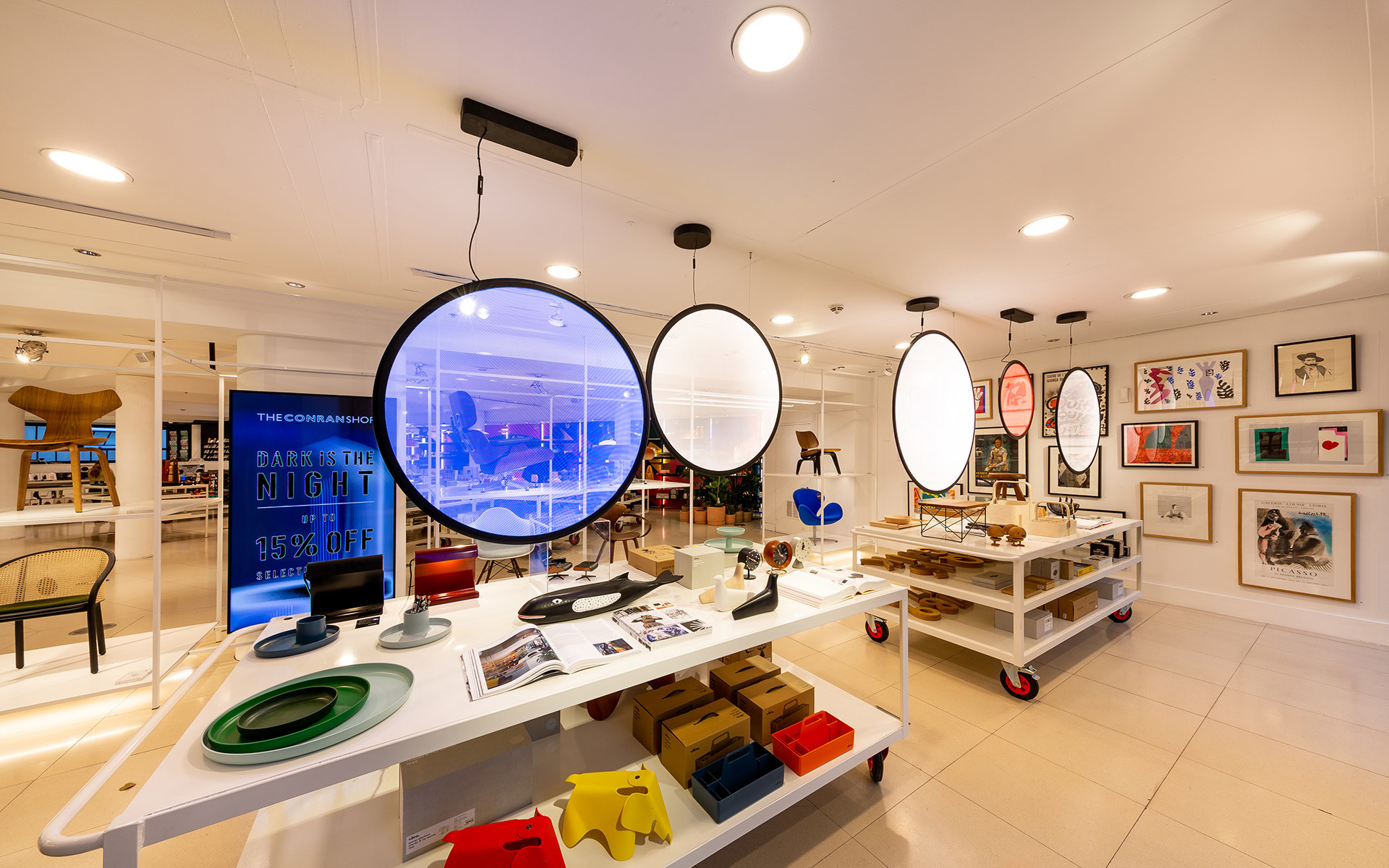 Spazio commerciale moderno con grandi lampade a sospensione circolari che illuminano tavoli espositivi con oggetti di design colorati.