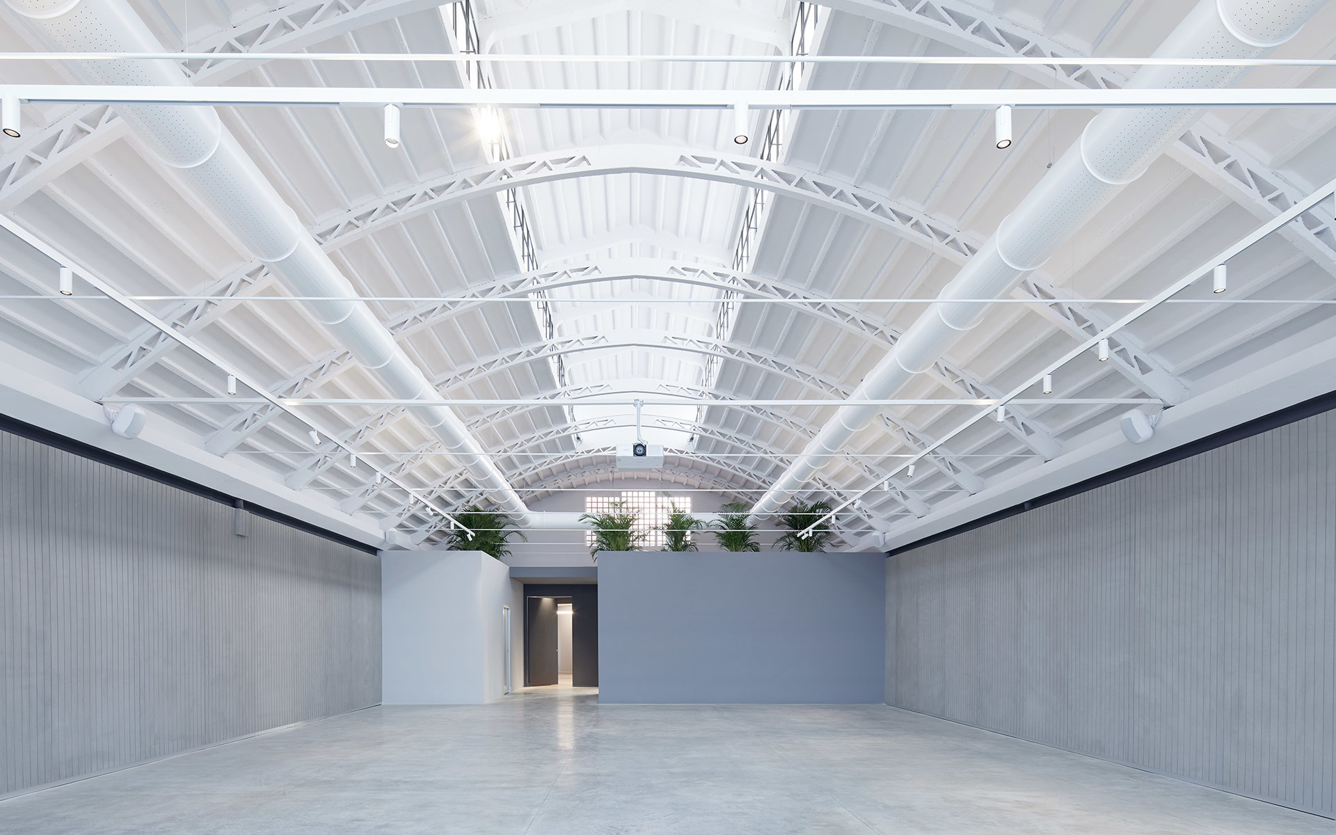 Spacious industrial hall with arched white steel roof structure, track lighting along beams, polished concrete floor, and plants along back wall.