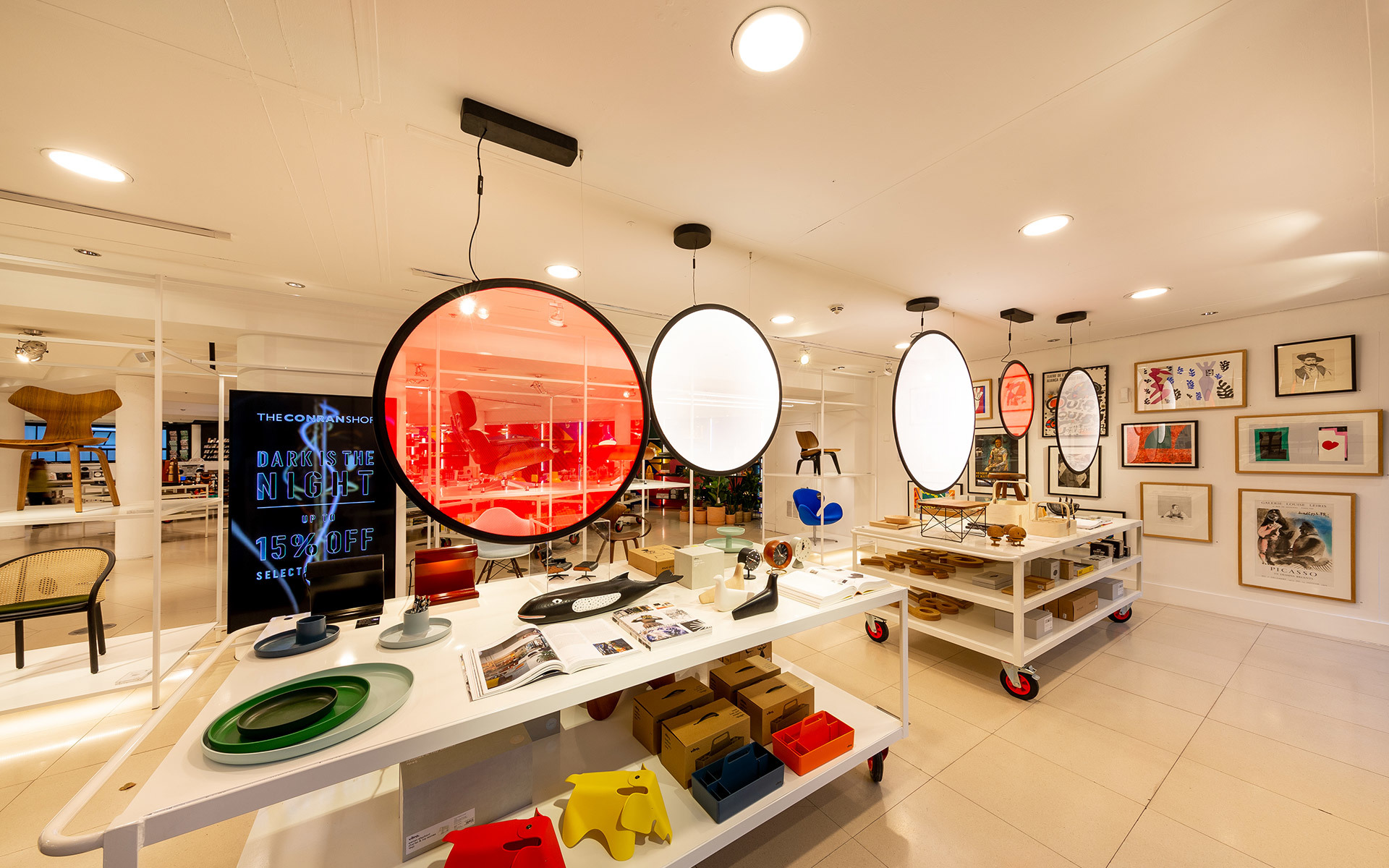 Negozio di design moderno con grandi lampade a sospensione ovali, tavoli espositivi bianchi con oggetti colorati e opere incorniciate alle pareti.