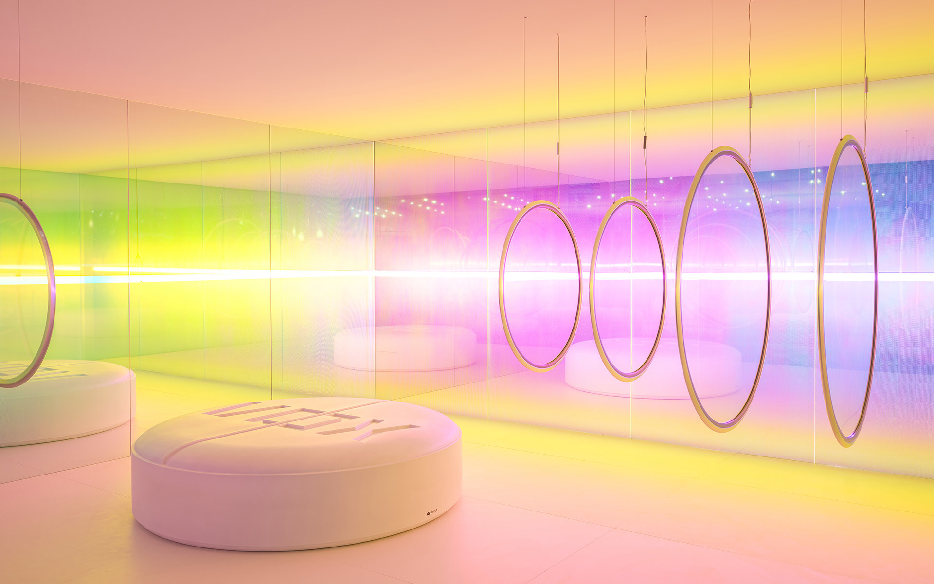 Stanza immersiva con lampade ad anello circolari a sospensione che proiettano vivaci gradienti arcobaleno su pareti specchiate e sedute curve bianche