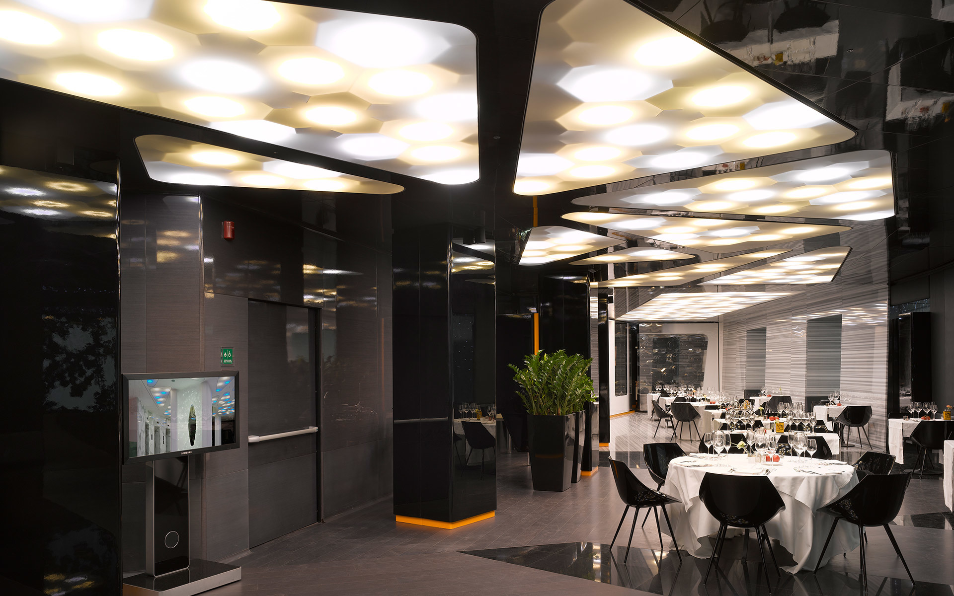 Interno di ristorante moderno con soffitto scuro e luminarie a incasso di forma organica disposte in gruppi, che illuminano i tavoli.
