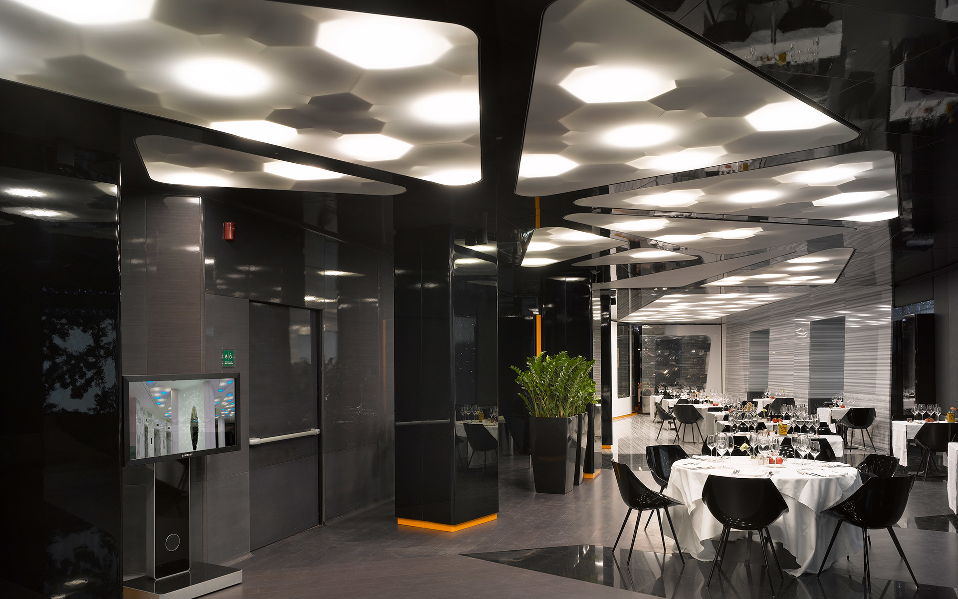 Interno di ristorante moderno con luci a soffitto esagonali che illuminano tavoli con tovaglie bianche in uno spazio elegante e scuro