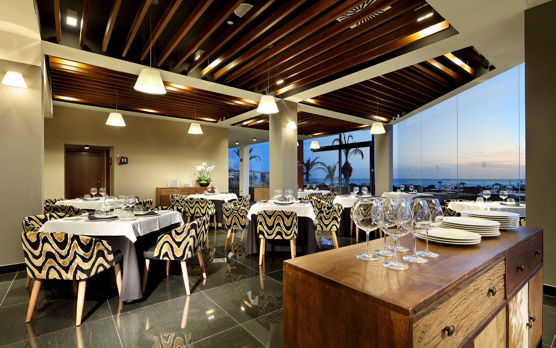 Elegant restaurant interior with pendant lights over white-clothed tables, bold patterned chairs, wood-slat ceiling, and ocean view at dusk.