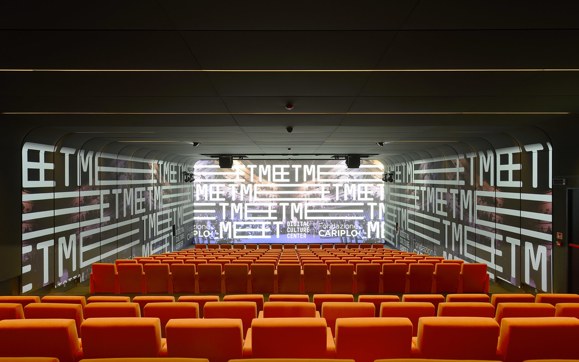 Modern auditorium with orange seats, ceiling-mounted spotlights, and large projection screen displaying 'TMEET' text pattern.