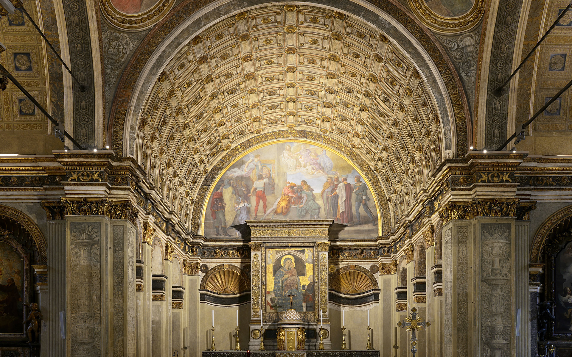 Ornate church apse with gilded coffered barrel vault, Renaissance frescoes, decorated pilasters, and a central altar with religious artwork.