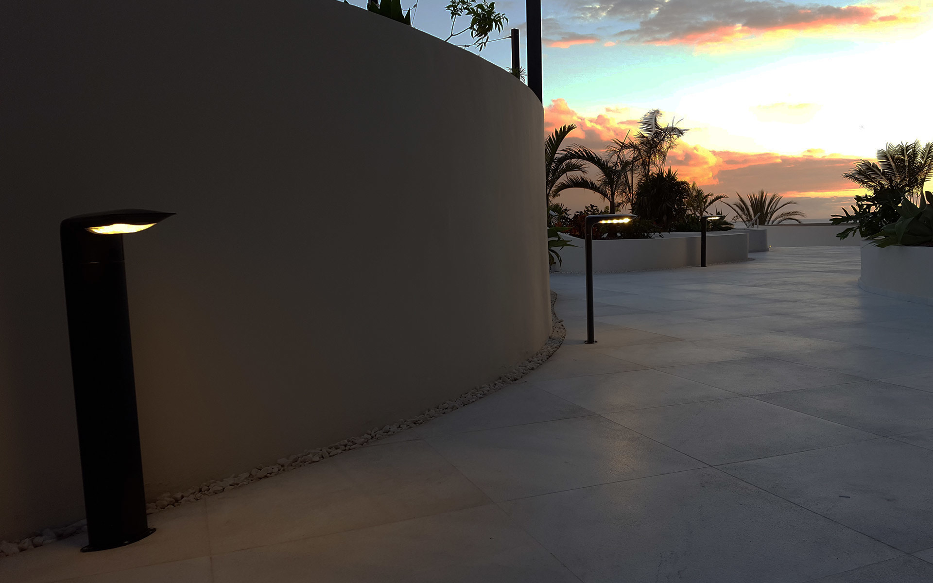 Dark bronze bollard light illuminating a terrace at sunset, with palm trees and ocean horizon in the background.