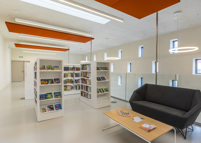Modern library reading area with Artemide circular pendant lamp, recessed linear lights, orange acoustic panels, white bookshelves, and a dark sofa
