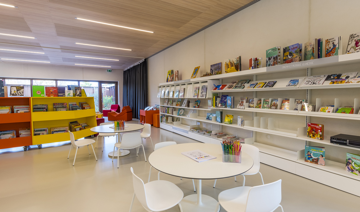 Bright children's library with Artemide linear ceiling lights, white shelves displaying colorful books, round tables, and white chairs.