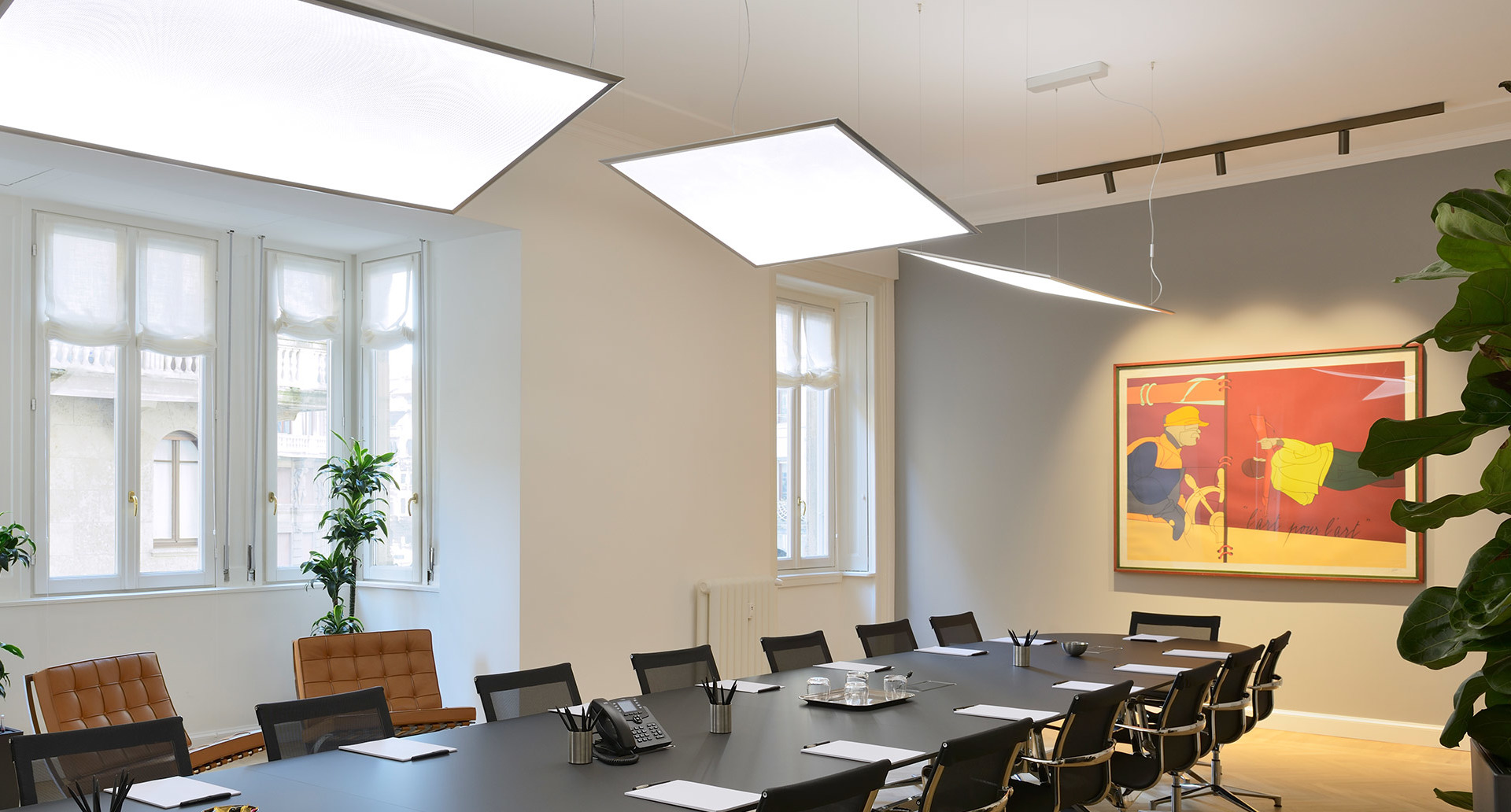 Modern conference room with large luminous ceiling panels and pendant linear lights illuminating a long dark meeting table with chairs.