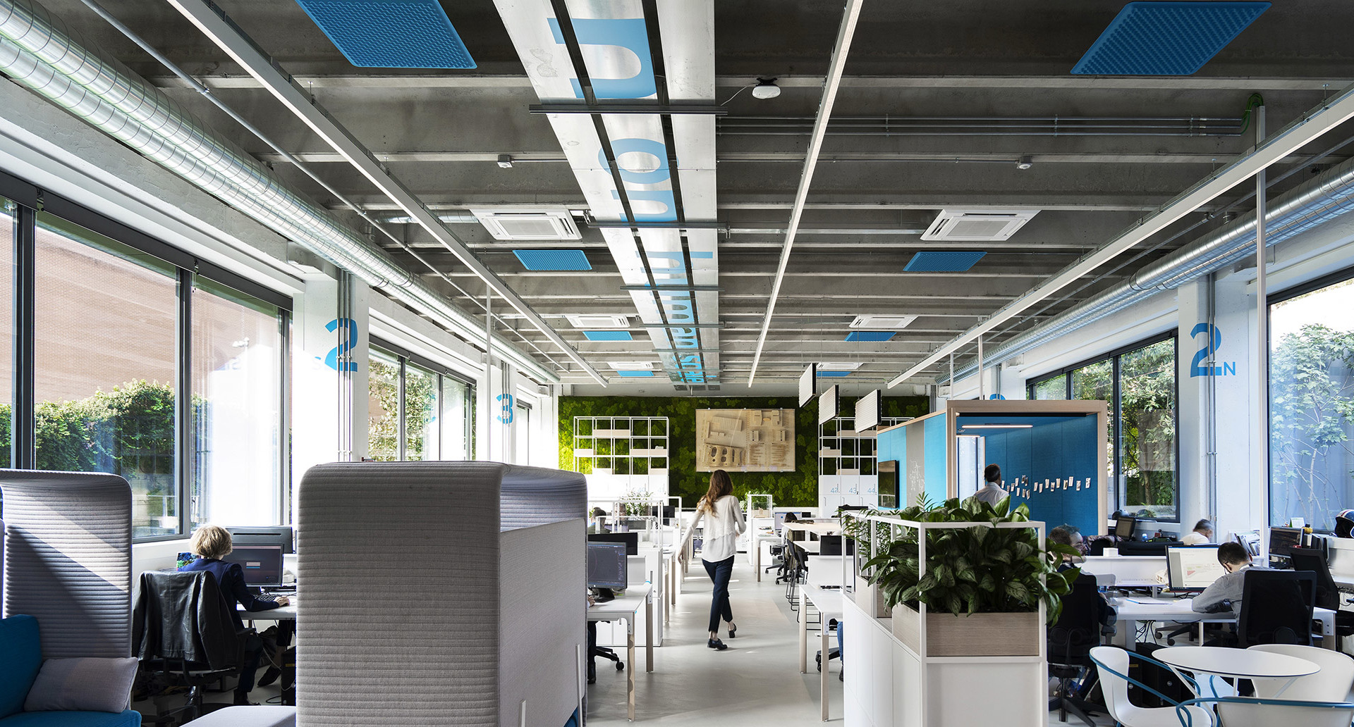 Ufficio open space moderno con pannelli luminosi Artemide a soffitto, postazioni di lavoro, cabine acustiche e piante verdi in luce naturale.