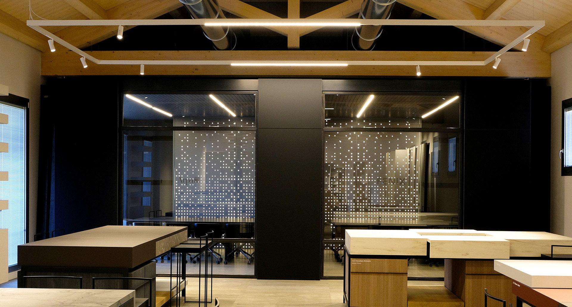 Modern workspace with Artemide track lights on wooden ceiling beams, illuminating tables and a dark partition wall with decorative perforated panels.
