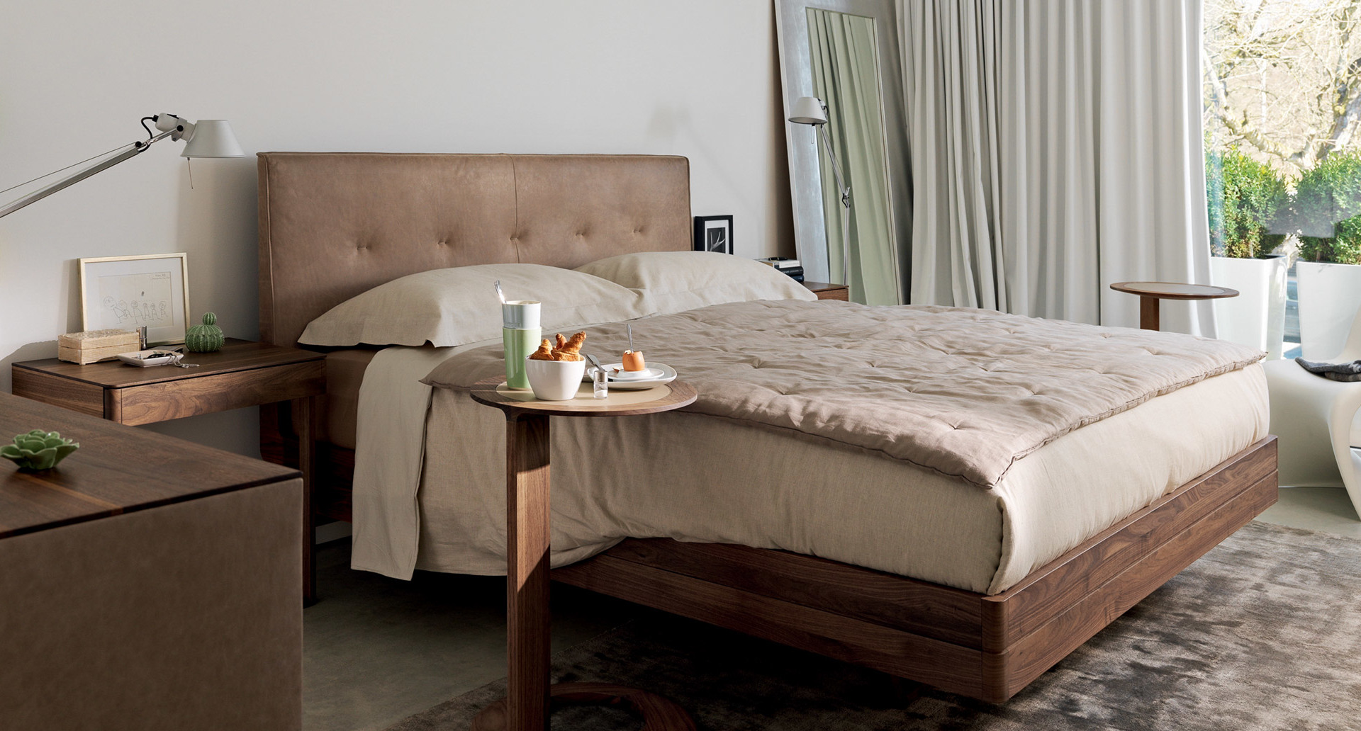Camera da letto elegante con letto in noce, testiera imbottita beige, biancheria neutra e lampada da tavolo regolabile Artemide sul comodino