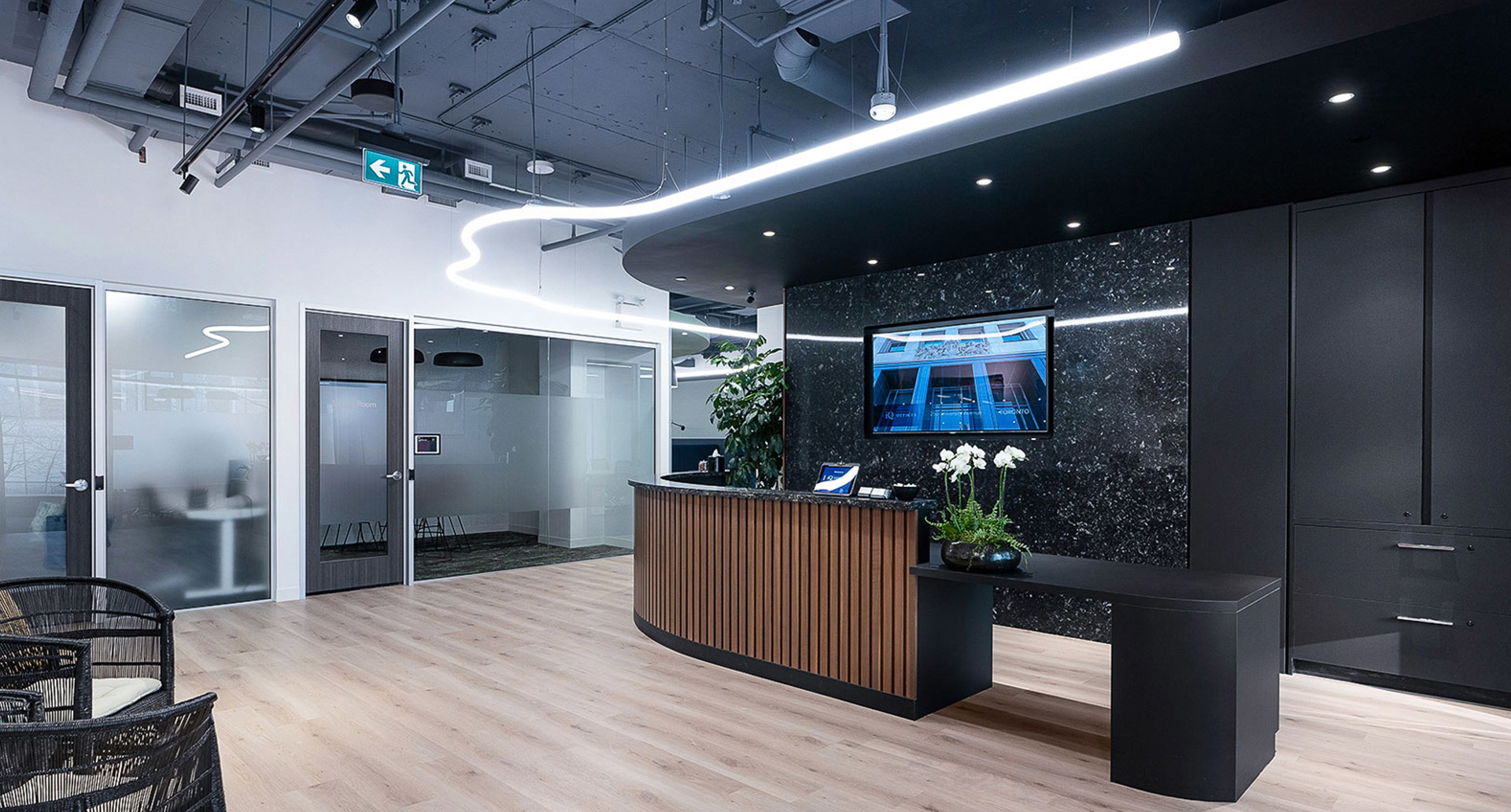 Reception ufficio moderna con luce lineare LED Artemide curva su soffitto scuro, scrivania in legno, parete in marmo e sale riunioni in vetro.