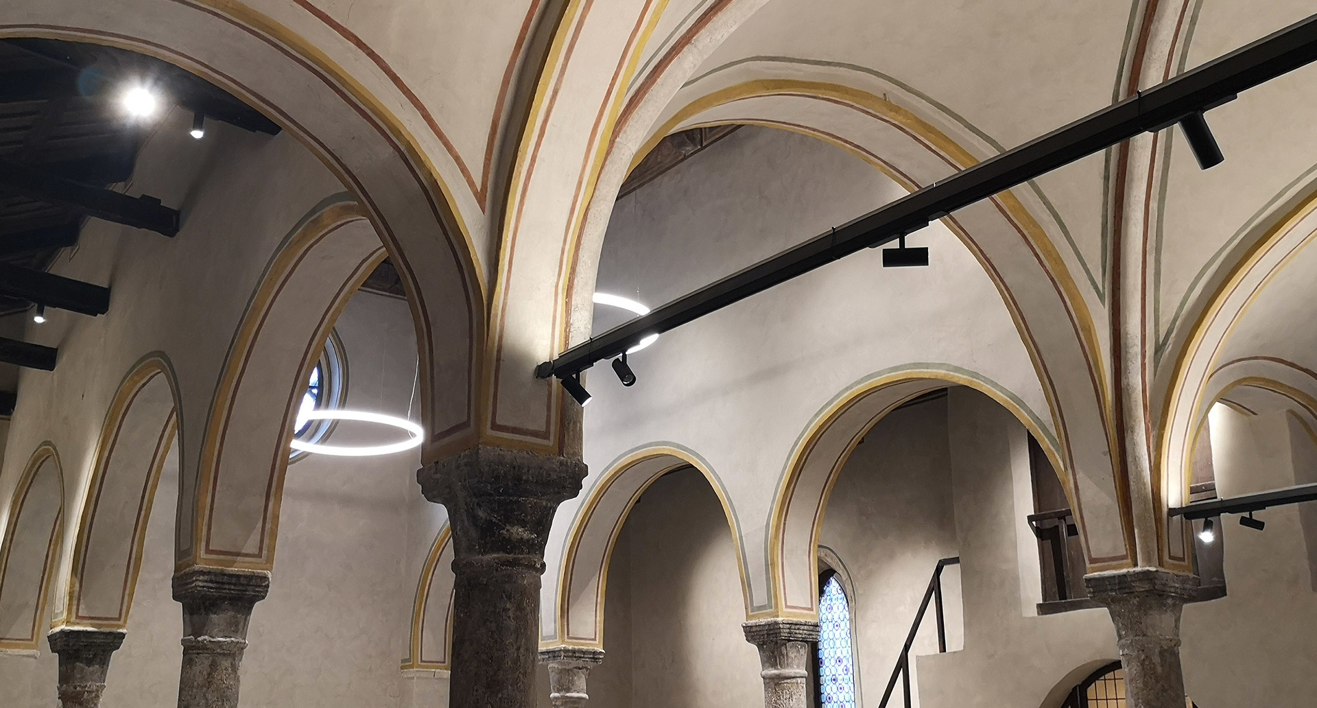 Sala con volte gotiche illuminata da sospensione circolare LED Artemide e faretti su binario nero che valorizzano archi e colonne in pietra