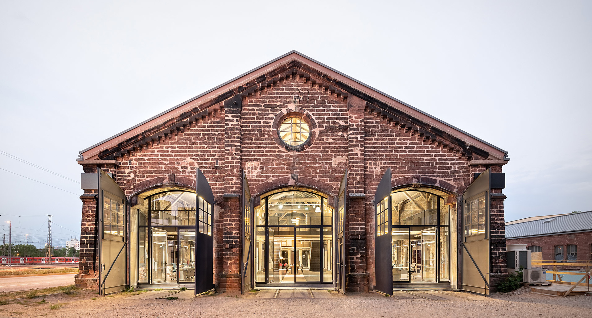 Edificio industriale storico in mattoni rossi ristrutturato con grandi finestre ad arco, interno illuminato caldo visibile al crepuscolo.
