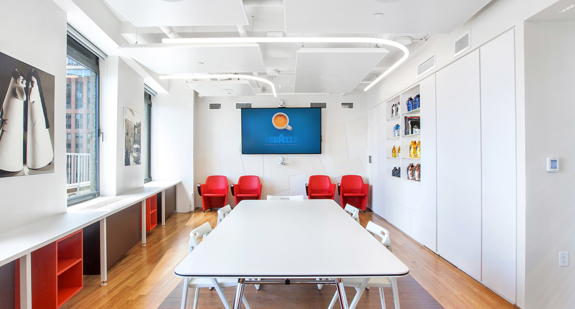 Sala conferenze moderna con luce a soffitto LED curva Artemide, tavolo bianco, sedie rosse e schermo di presentazione su parete bianca.