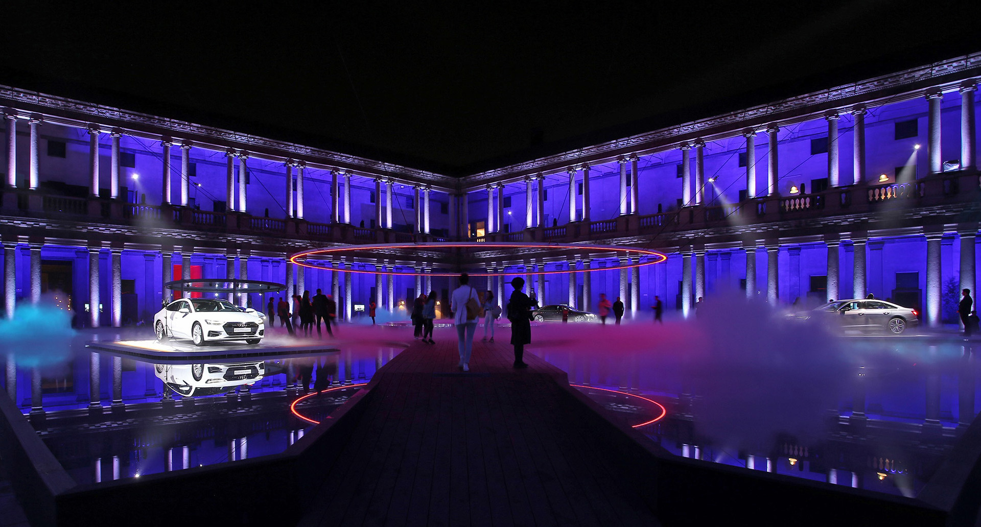 Grande cortile neoclassico illuminato in blu intenso con colonne illuminate, due auto bianche in esposizione, installazione luminosa circolare rossa ed effetti nebbia notturni