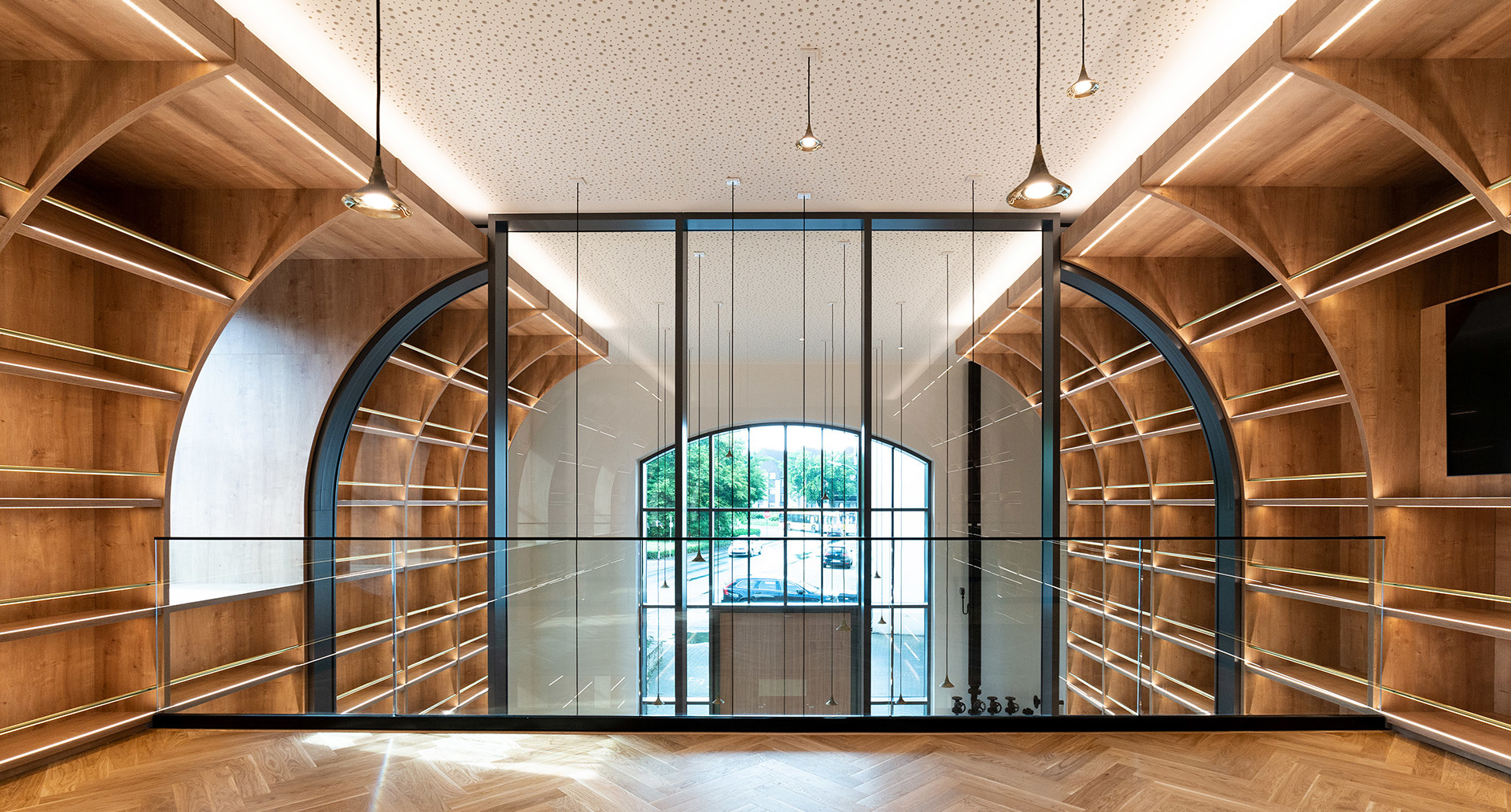 Interno elegante con scaffalature in legno caldo, divisorio in vetro ad arco, pavimento a spina di pesce e lampade a sospensione Artemide dal soffitto forato.
