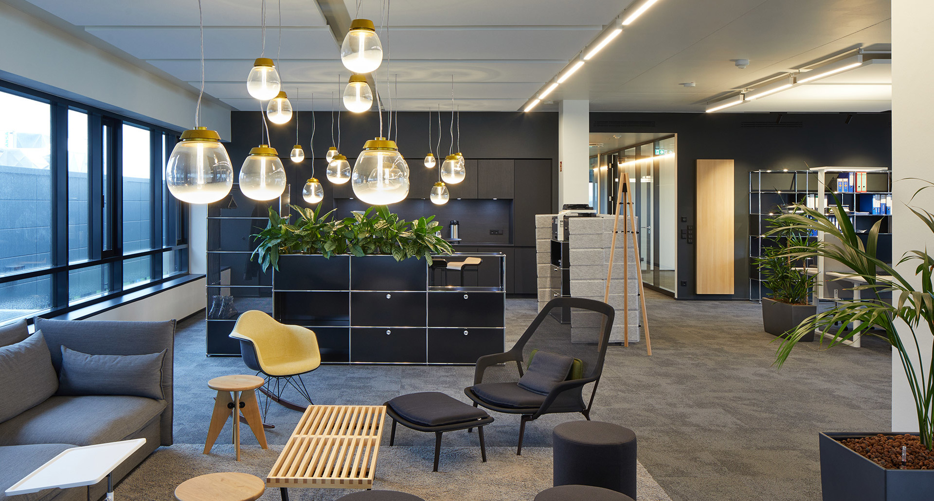 Lounge ufficio moderno con lampade a sospensione Artemide con globi in vetro e sommità dorata, divani grigi e piante verdi.