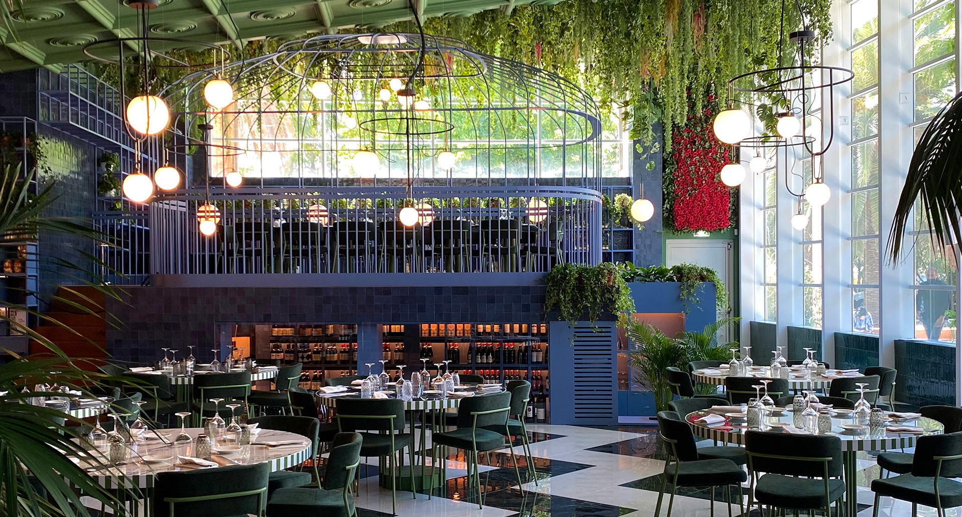Elegant restaurant interior with Artemide globe pendant lights and circular chandelier amid lush hanging greenery, green chairs, and checkered floor.