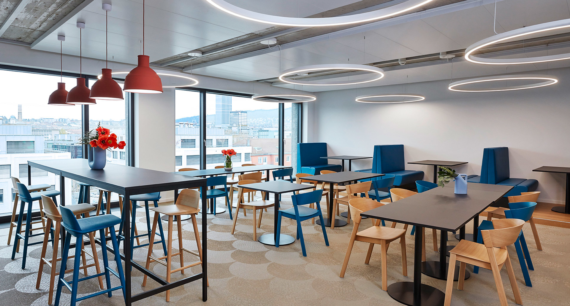 Mensa ufficio moderna con anelli LED circolari Artemide a soffitto, lampade a sospensione rosse, sedie blu e legno, ampie finestre con vista città