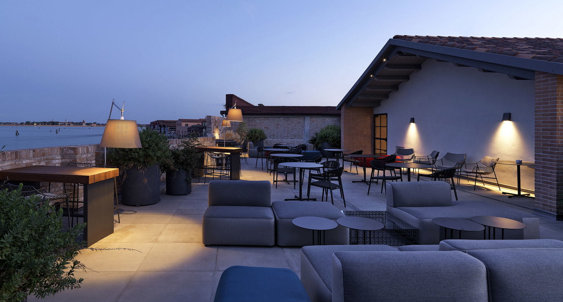 Terrazza sul tetto al crepuscolo con lampade da terra e da parete Artemide che illuminano zone lounge e ristoro con vista sull'acqua