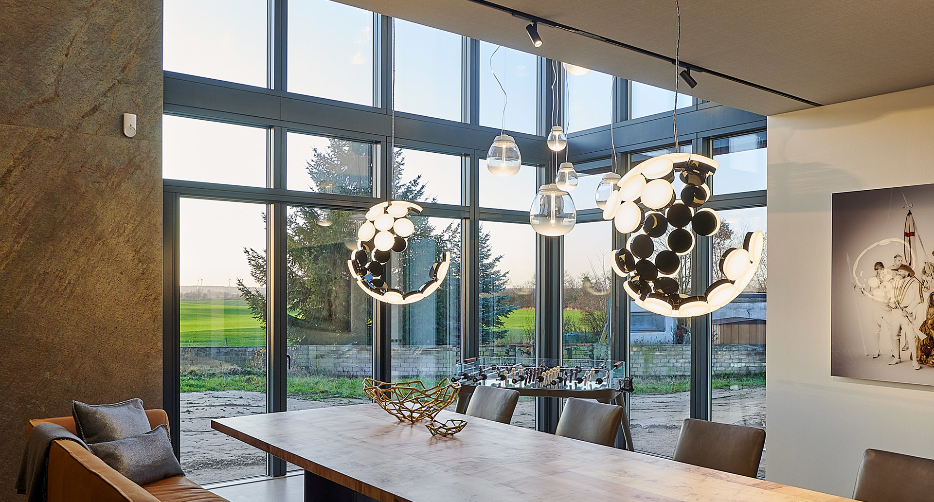 Sala da pranzo con grandi finestre e tre lampade a sospensione Artemide con cluster circolari LED bianco-neri sopra un tavolo in legno.