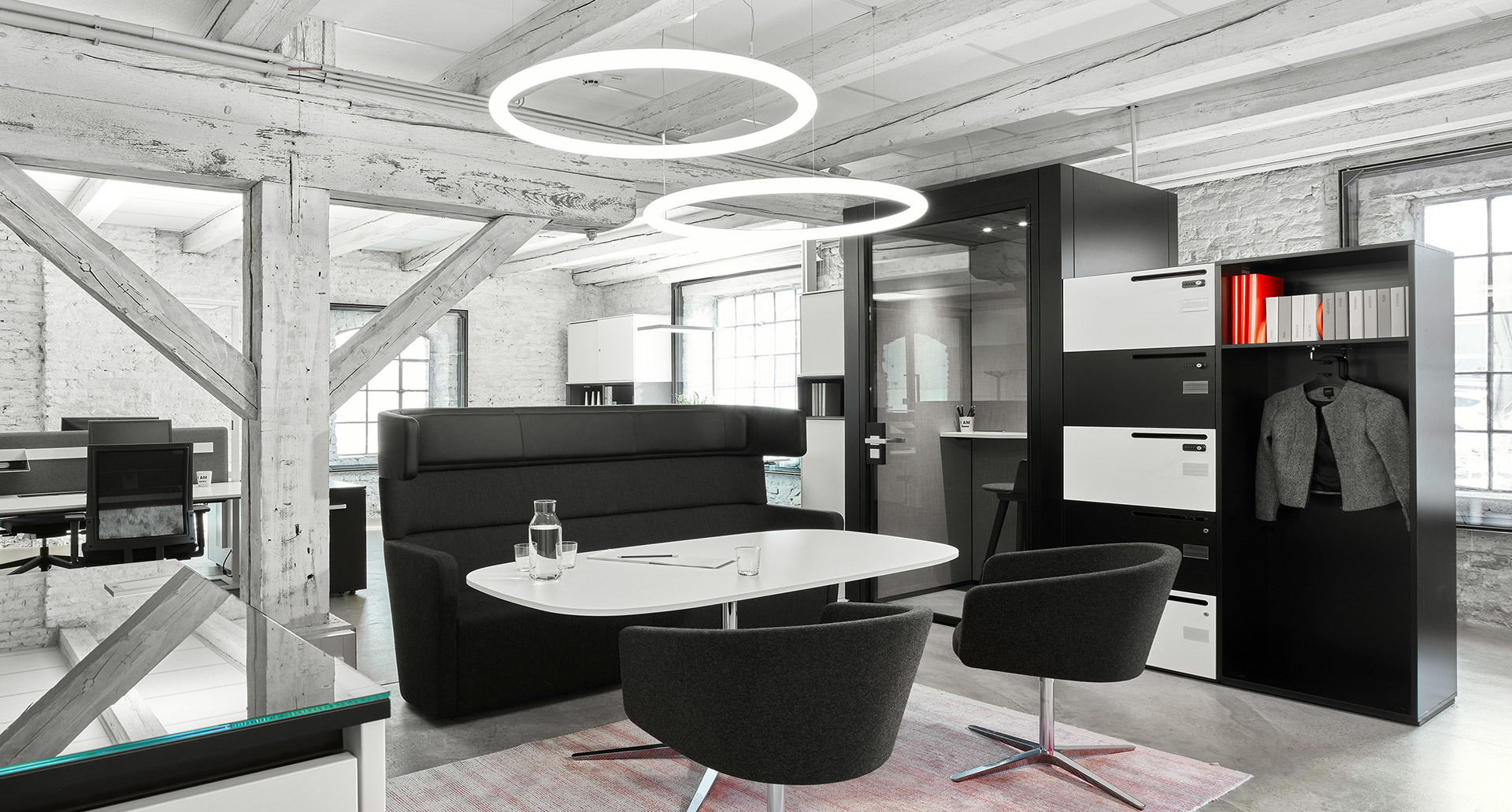 Lounge ufficio moderno con due lampade a sospensione ad anello circolare Artemide che illuminano sedute nere, tavolo ovale e pareti in mattoni industriali.