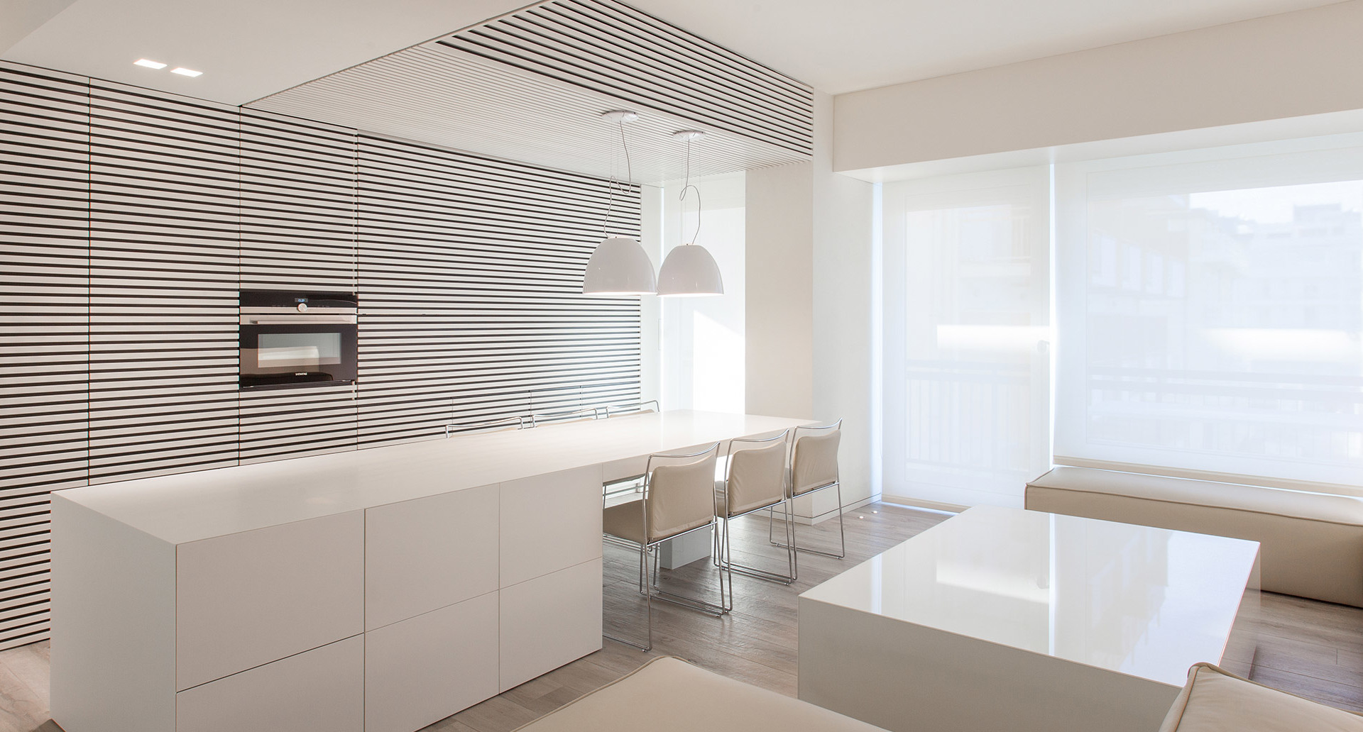Modern white kitchen with pendant lamp over island, dining area with chairs, striped wall panel, and large window with sheer blind